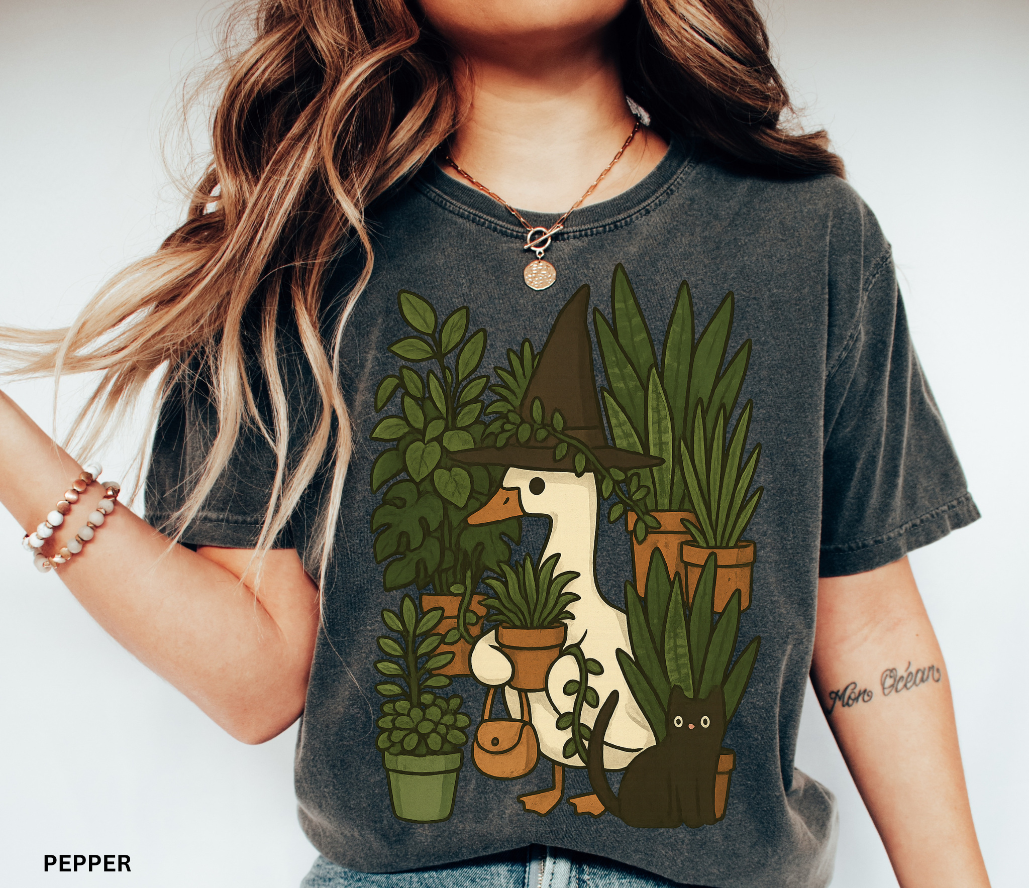 Goose wearing a witch hat surrounded by plants with a black cat companion — cozy cottagecore witchy design on Comfort Colors shirt.