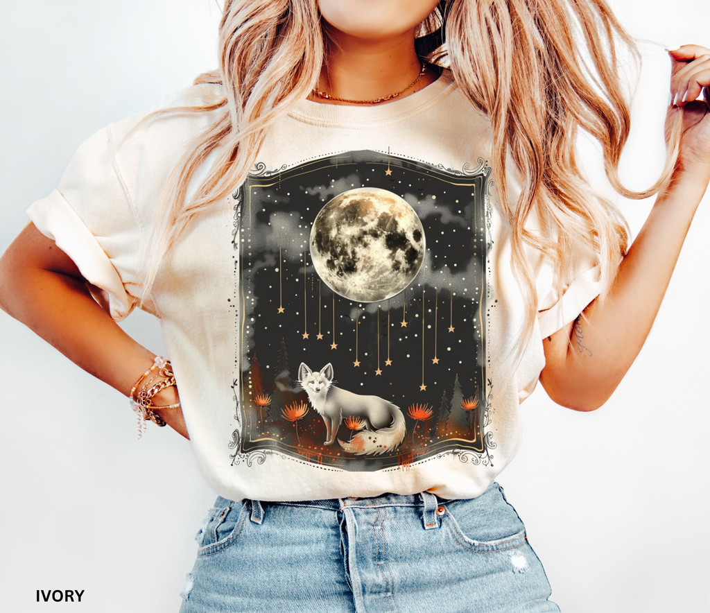 A mystical fox standing beneath a glowing full moon surrounded by stars and forest flowers on a celestial-themed shirt.