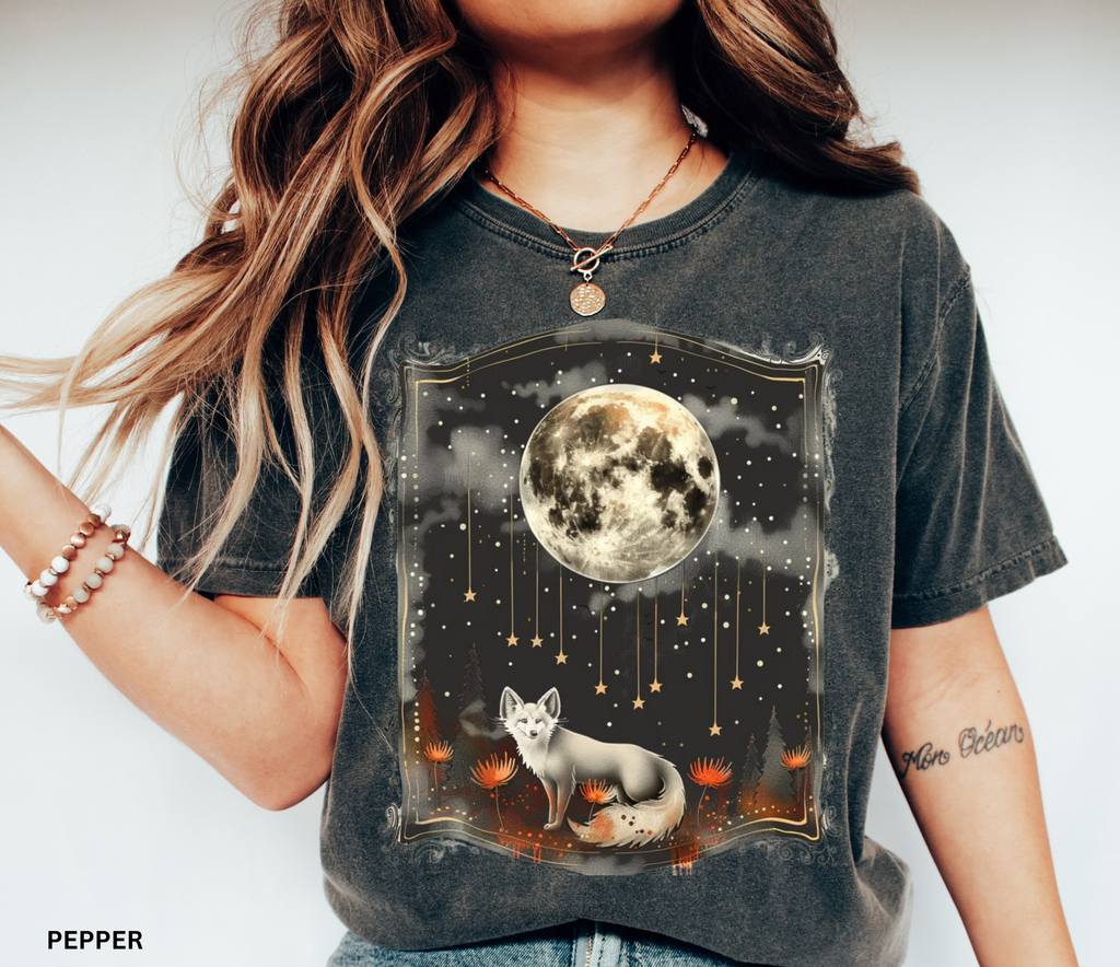 A mystical fox standing beneath a glowing full moon surrounded by stars and forest flowers on a celestial-themed shirt.