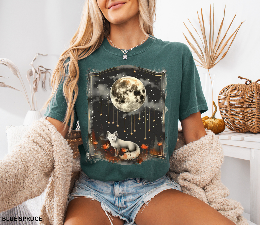 A mystical fox standing beneath a glowing full moon surrounded by stars and forest flowers on a celestial-themed shirt.