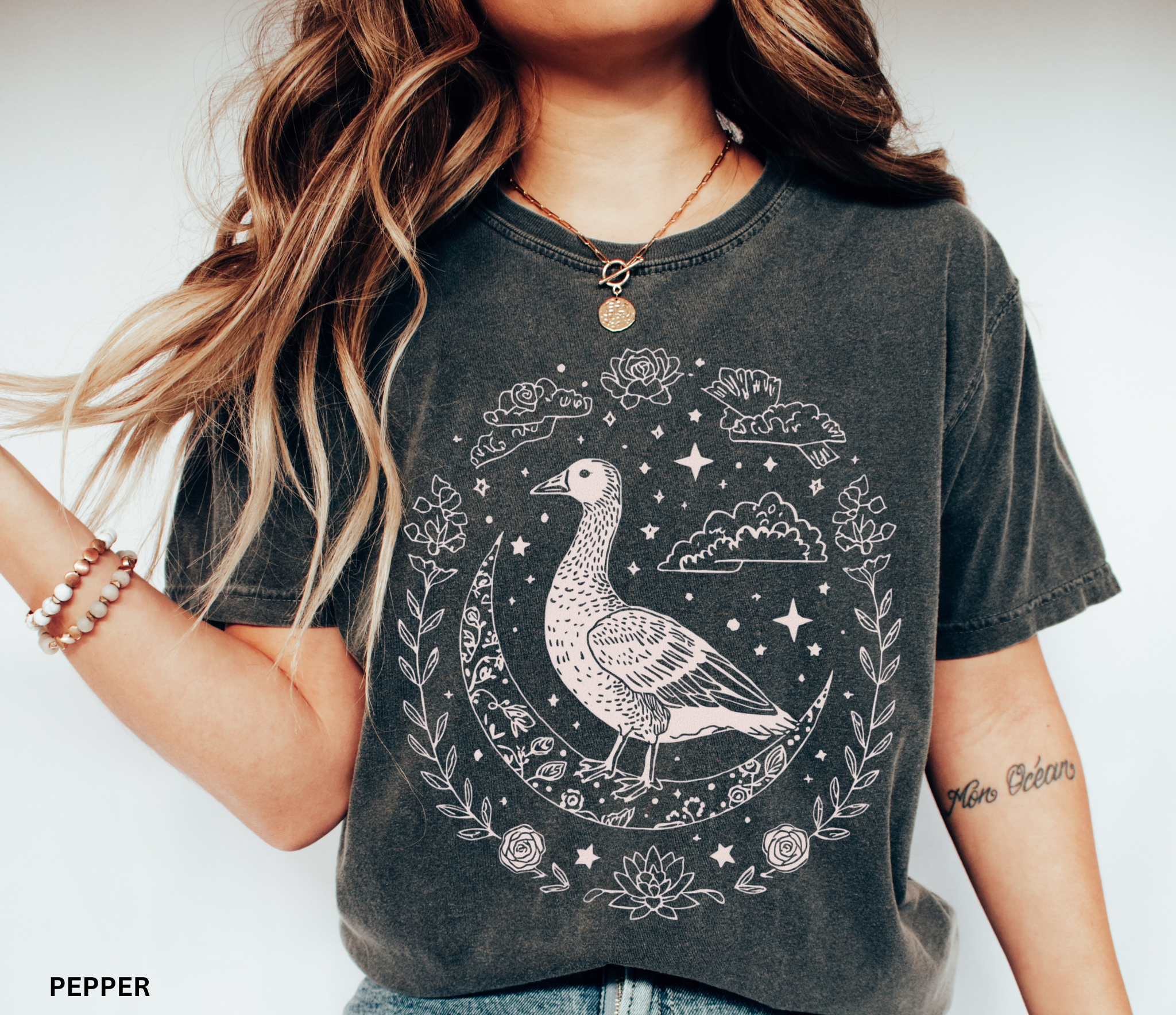 A mystical goose standing on a crescent moon surrounded by stars and floral borders, printed on a soft Comfort Colors witchy cottagecore shirt.