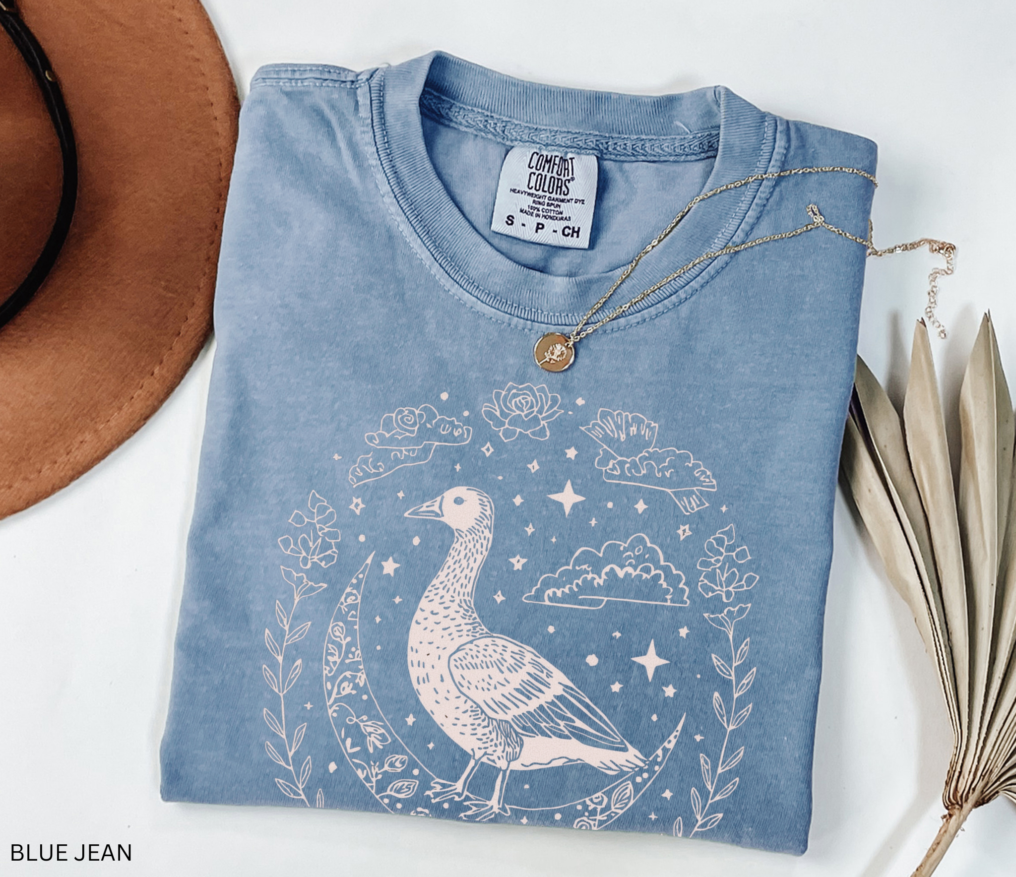 A mystical goose standing on a crescent moon surrounded by stars and floral borders, printed on a soft Comfort Colors witchy cottagecore shirt.