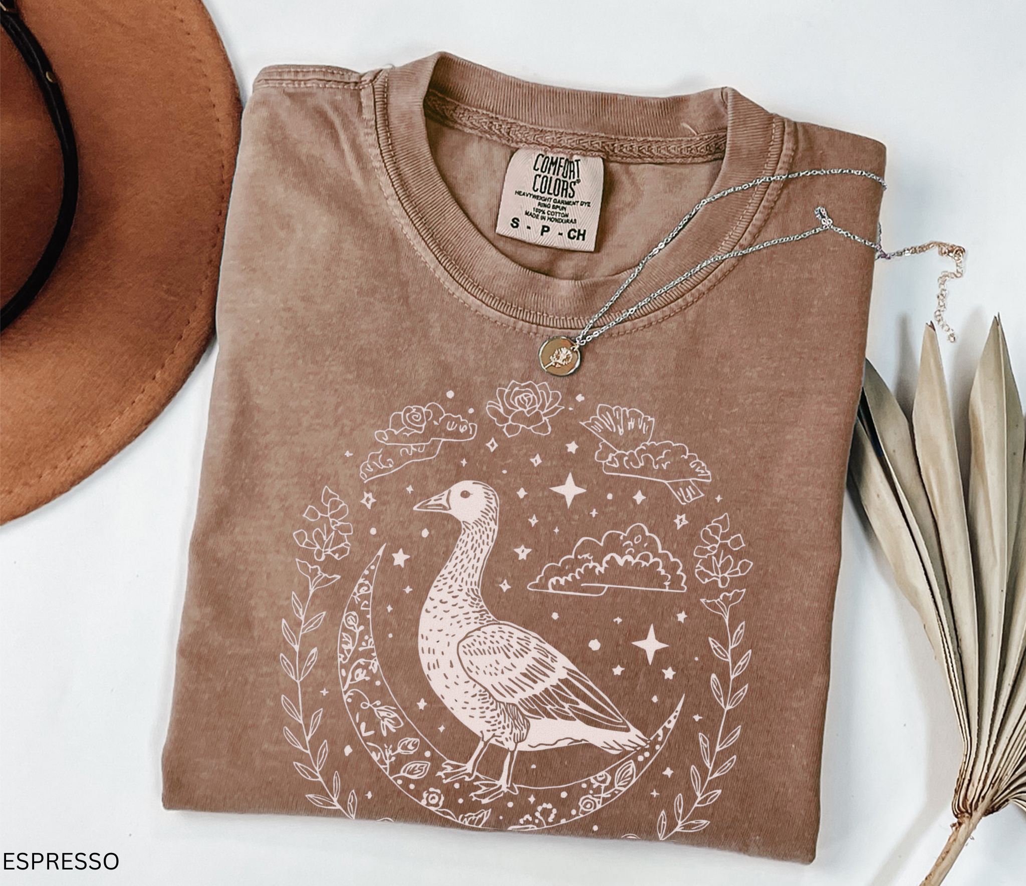 A mystical goose standing on a crescent moon surrounded by stars and floral borders, printed on a soft Comfort Colors witchy cottagecore shirt.