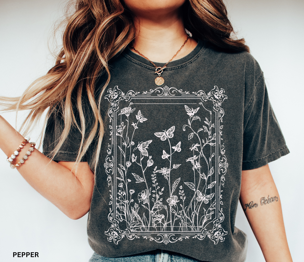 An ornate Victorian-style frame encasing hand-drawn butterflies and wildflowers on a whimsigoth-inspired cottagecore shirt.