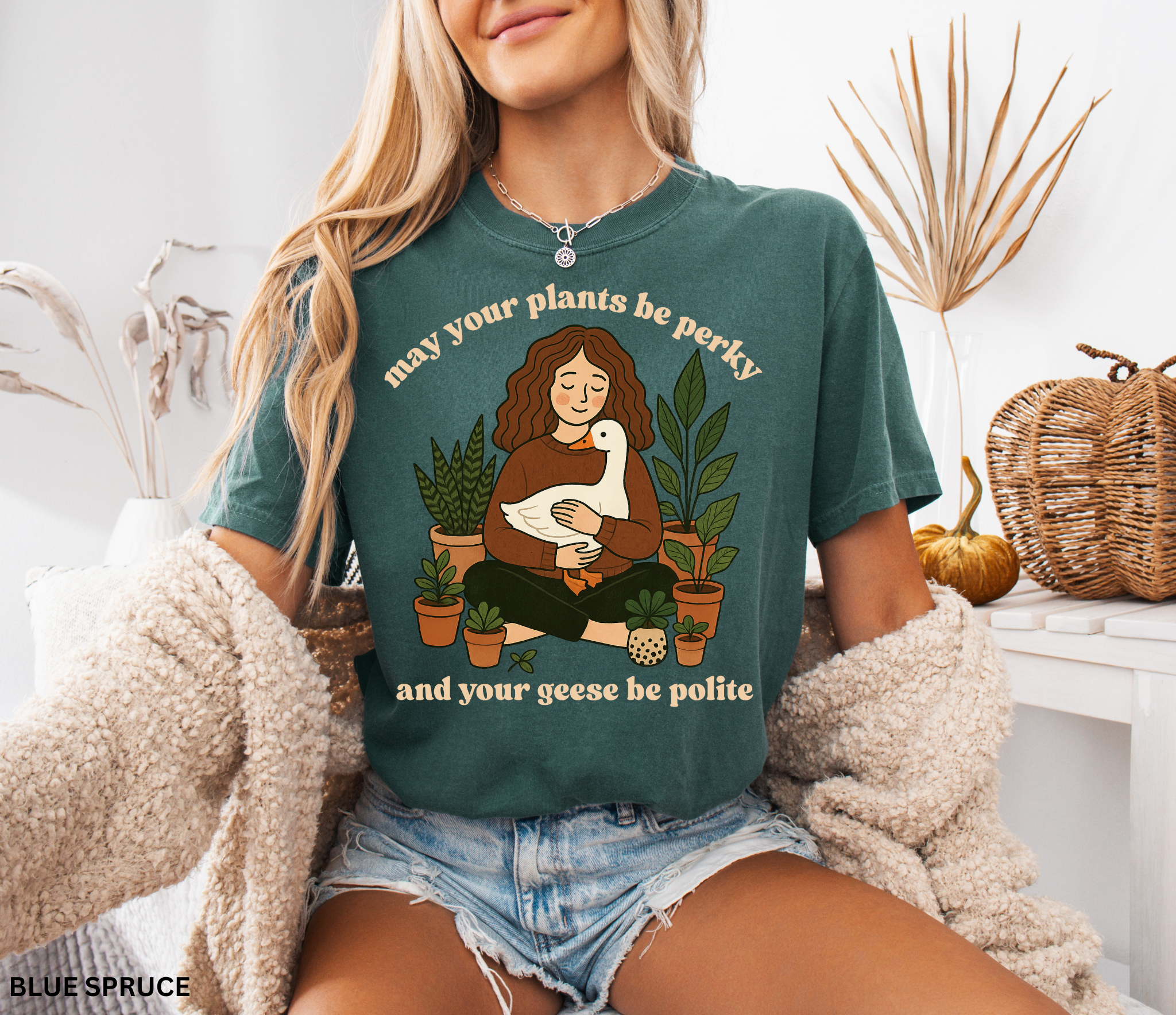 A cozy woman holding a goose surrounded by potted plants with the text “May your plants be perky and your geese be polite” printed on a Comfort Colors cottagecore shirt.