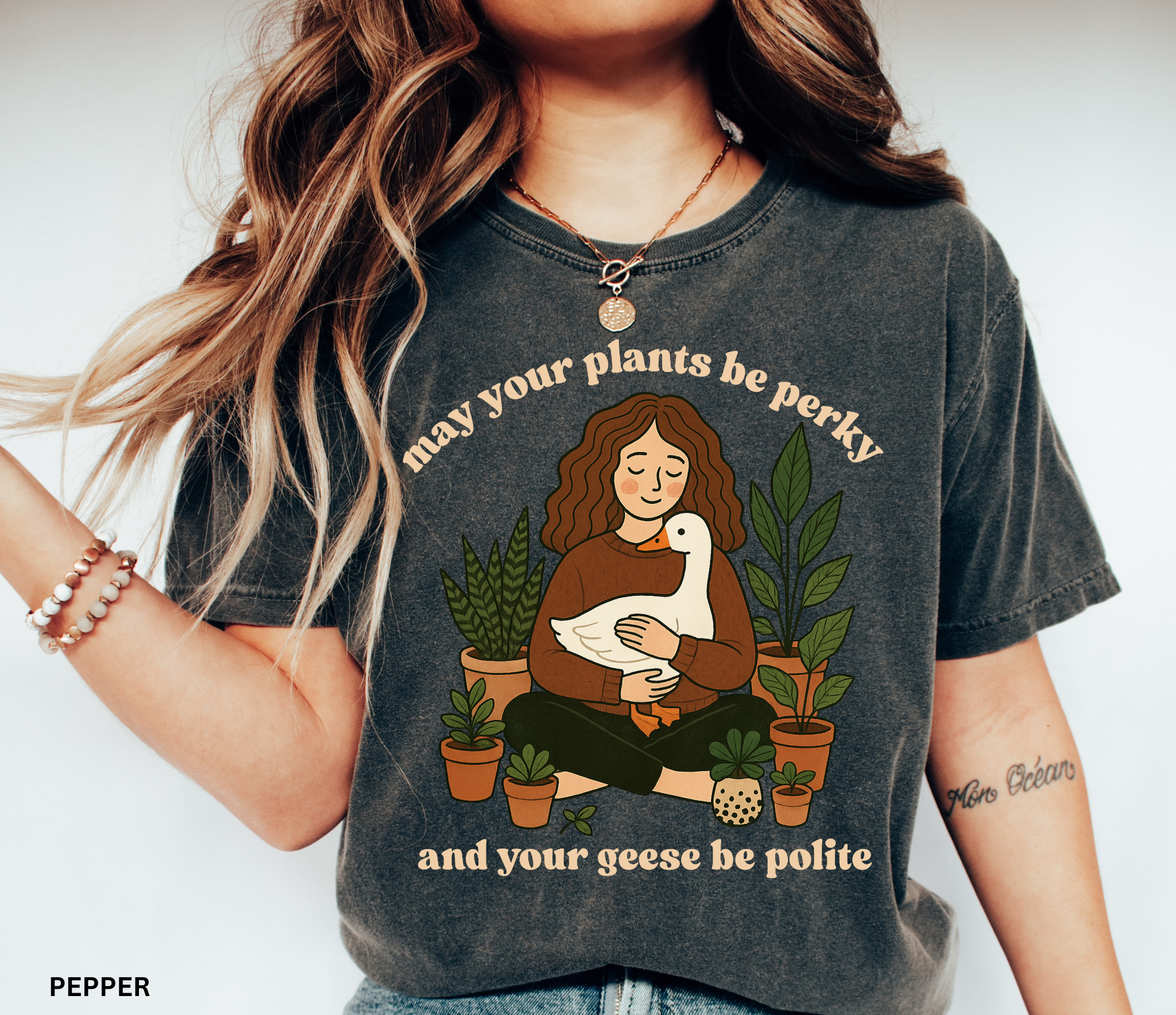 A cozy woman holding a goose surrounded by potted plants with the text “May your plants be perky and your geese be polite” printed on a Comfort Colors cottagecore shirt.