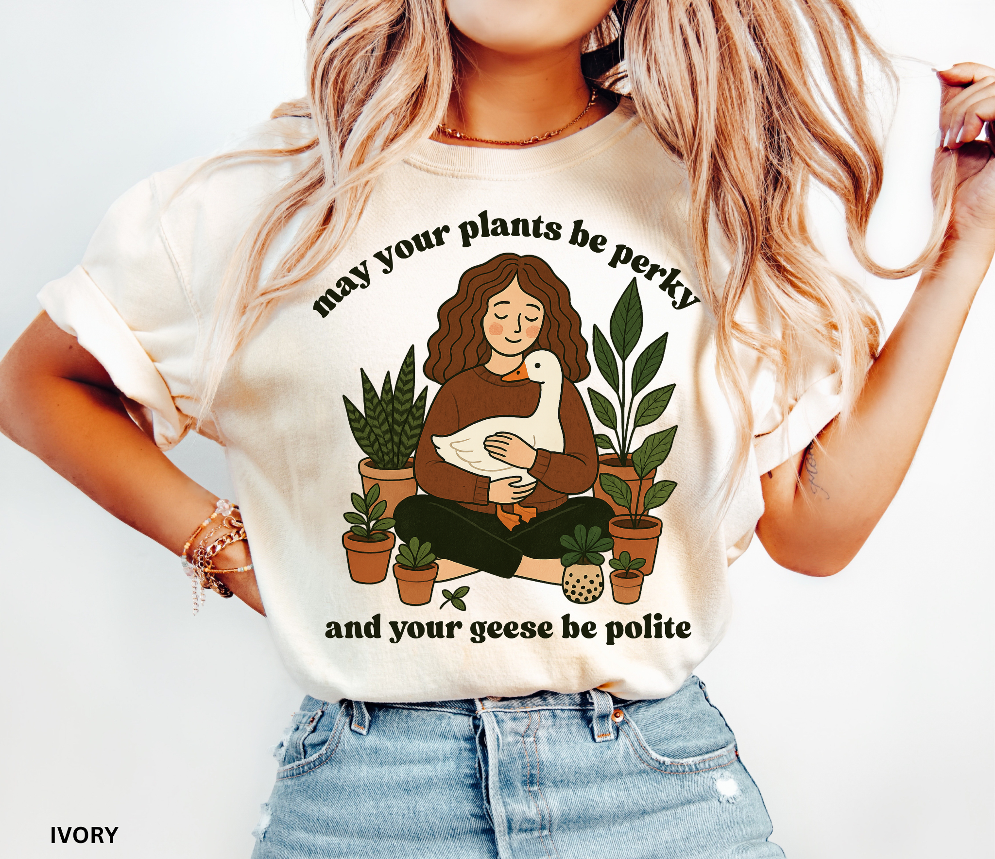 A cozy woman holding a goose surrounded by potted plants with the text “May your plants be perky and your geese be polite” printed on a Comfort Colors cottagecore shirt.