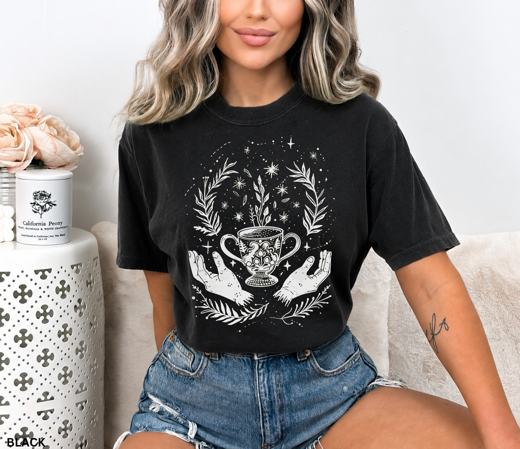Black Comfort Colors style t-shirt featuring a mystical tea chalice held between open hands, surrounded by stars, botanical leaves, and celestial witchy elements in a cottagecore tea witch aesthetic.