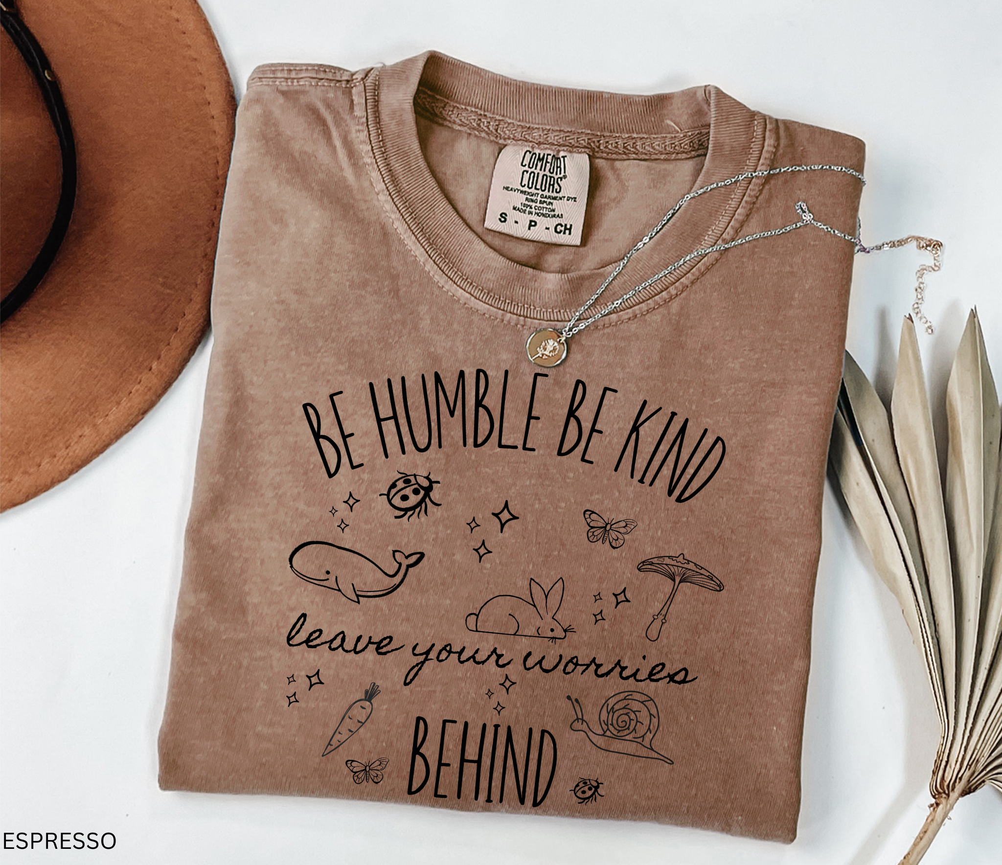  shirt featuring the quote "Be Humble Be Kind Leave Your Worries Behind" surrounded by whimsical illustrations including a rabbit, whale, mushroom, snail, butterflies, and ladybugs in a cottagecore style.