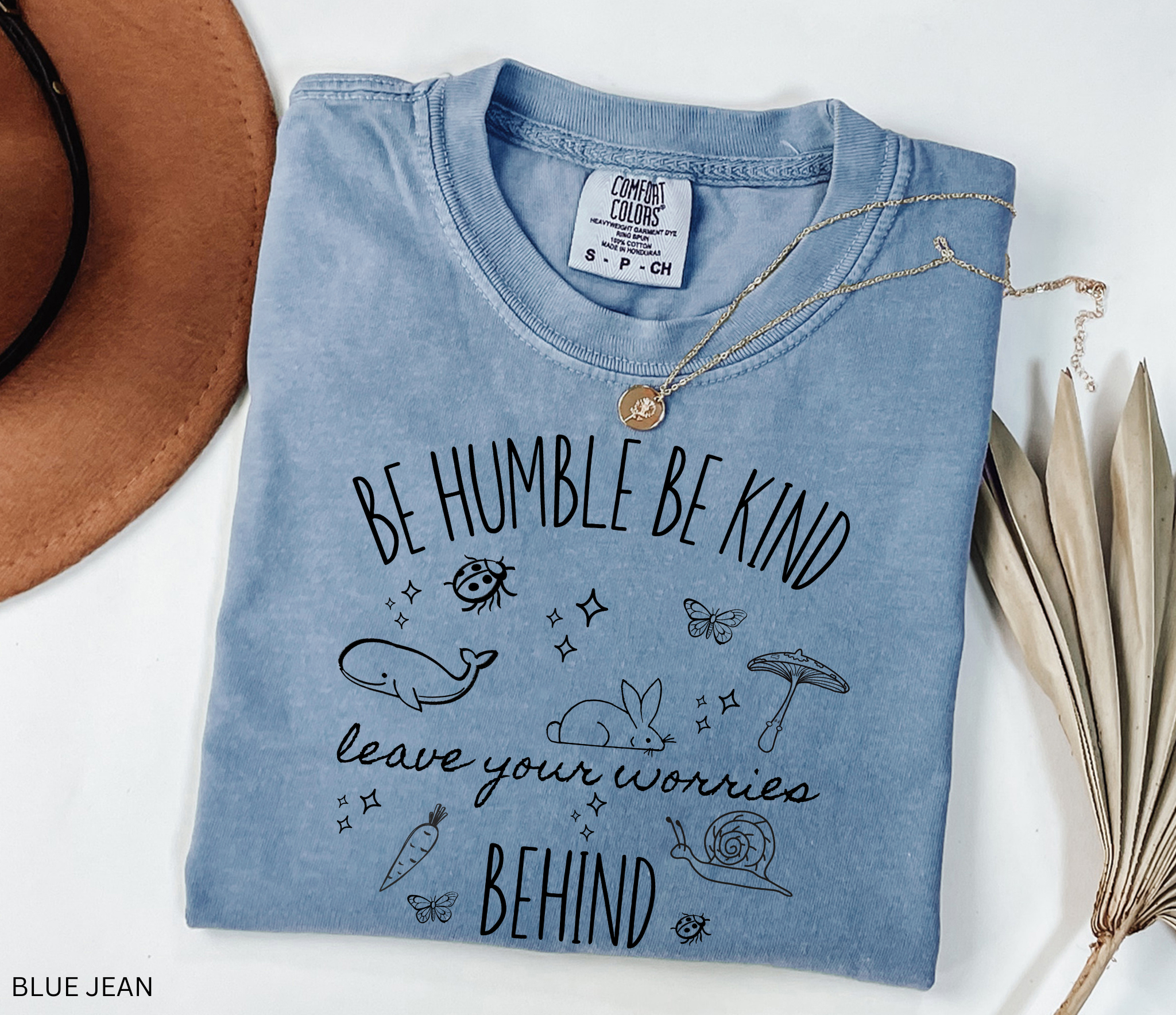 Comfort Colors shirt featuring the quote "Be Humble Be Kind Leave Your Worries Behind" surrounded by whimsical illustrations including a rabbit, whale, mushroom, snail, butterflies, and ladybugs in a cottagecore style.