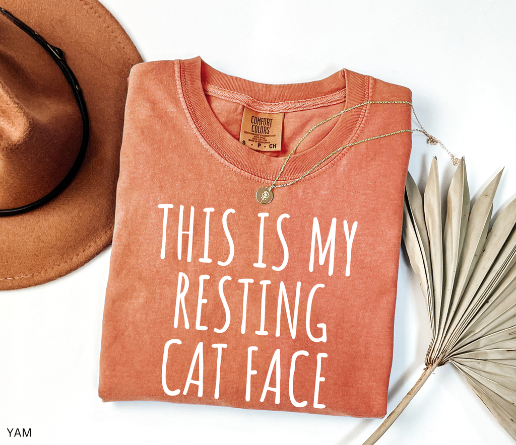Folded orange t-shirt with 'This is my resting cat face' text, next to a brown hat and dried plant on a white background.