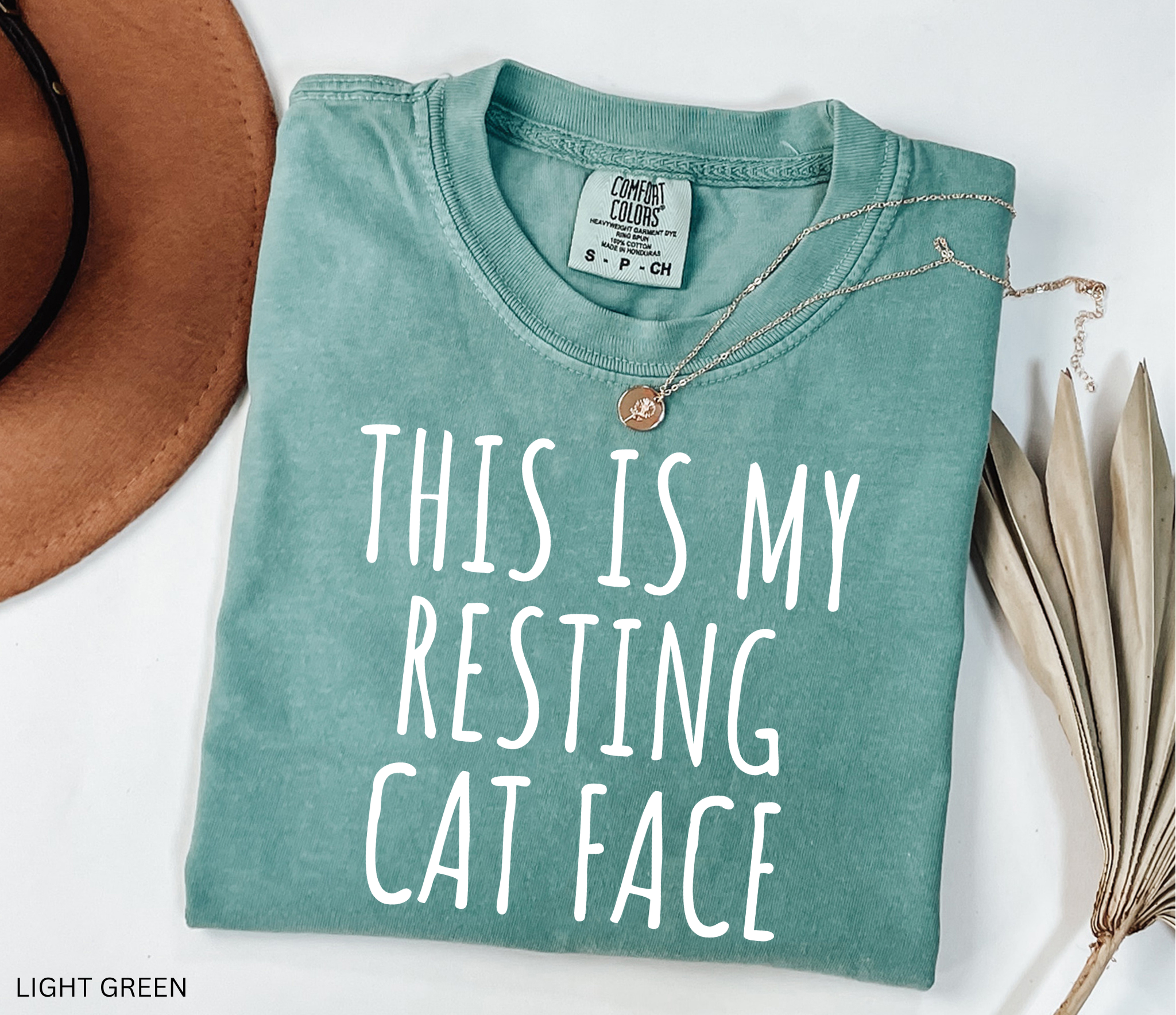 Light green t-shirt with 'This is my resting cat face' text, placed on a white surface with a hat and dried leaves.