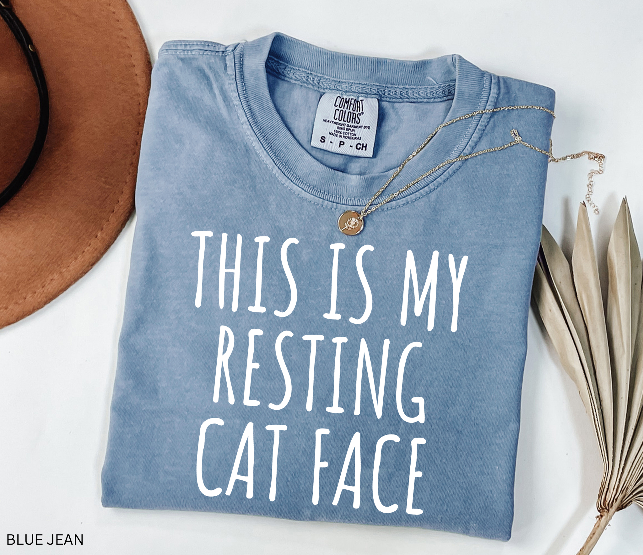 Folded blue shirt with 'This is my resting cat face' text on a white surface with a hat and plant.