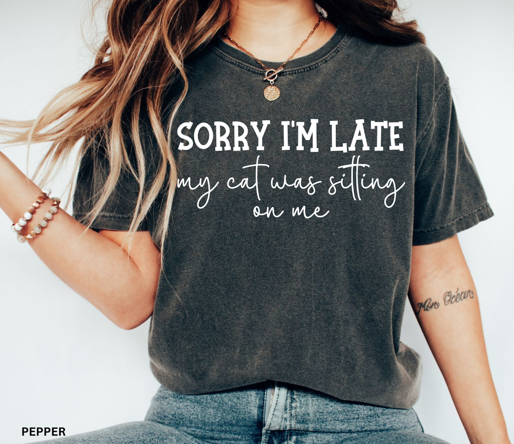 Funny cat lover graphic t-shirt featuring the quote "Sorry I'm Late My Cat Was Sitting on Me." A humorous gift for cat moms, cat dads, and anyone owned by a sleepy cat.