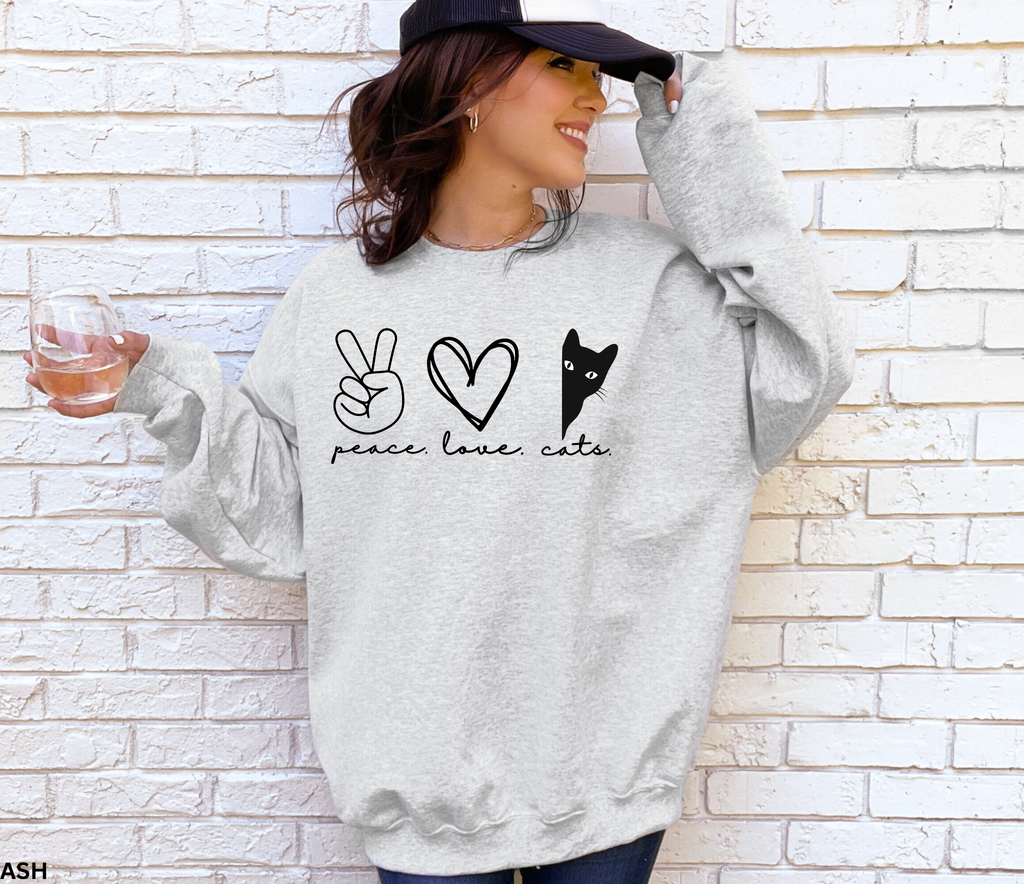 Peace Love Cats Gildan 18000 sweatshirt in sand featuring a peace sign, heart, and peeking black cat graphic, folded flat on a wood background.