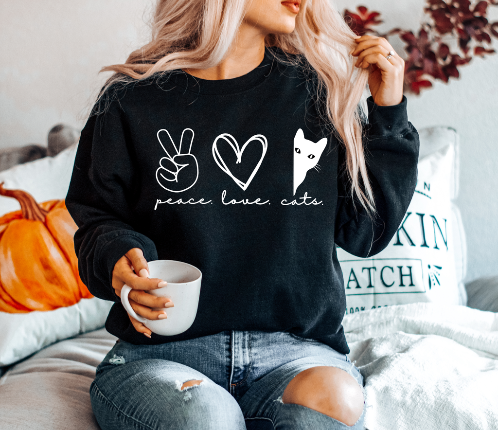 Woman wearing a black Peace Love Cats sweatshirt featuring a white peace sign, heart, and peeking black cat graphic while holding a coffee mug.