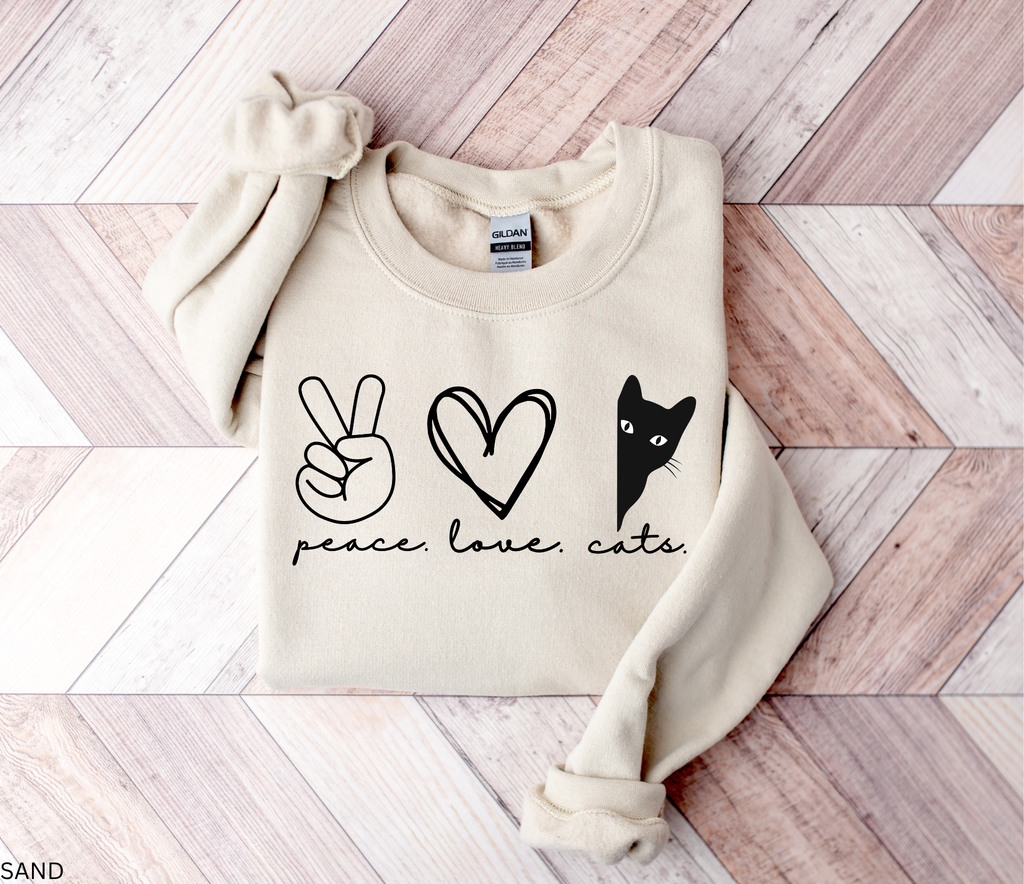 Peace Love Cats Gildan 18000 sweatshirt in sand featuring a peace sign, heart, and peeking black cat graphic, folded flat on a wood background.