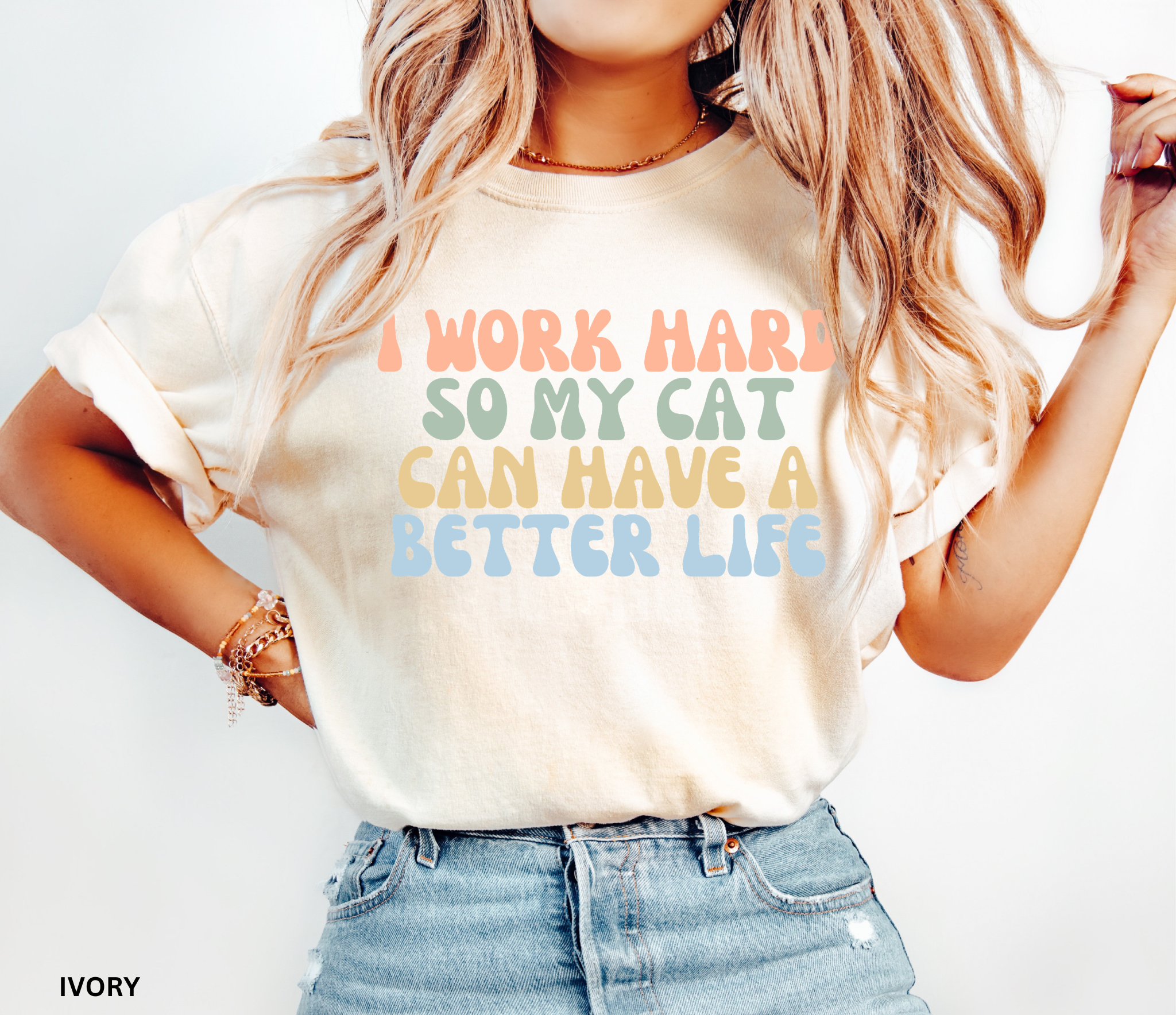 A woman wearing a light ivory graphic t-shirt with the text 'I WORK HARD SO MY CAT CAN HAVE A BETTER LIFE' in pastel orange, green, yellow, and blue lettering, paired with light blue denim shorts.