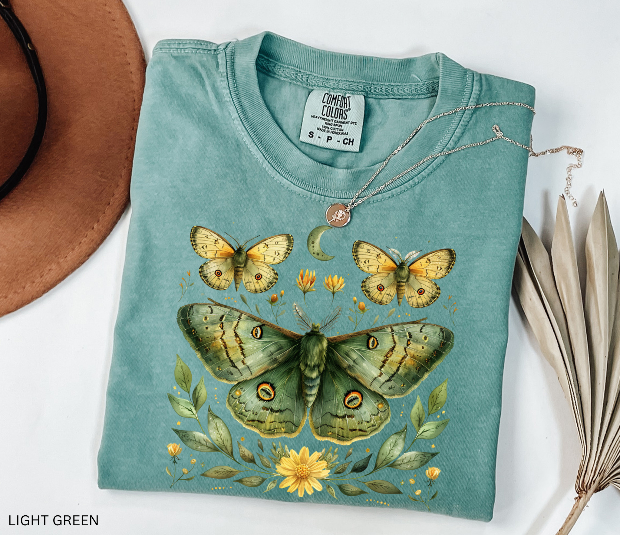 Comfort Colors graphic t-shirt featuring a detailed green luna moth surrounded by wildflowers, leaves, smaller moths, and a crescent moon in a vintage botanical cottagecore illustration