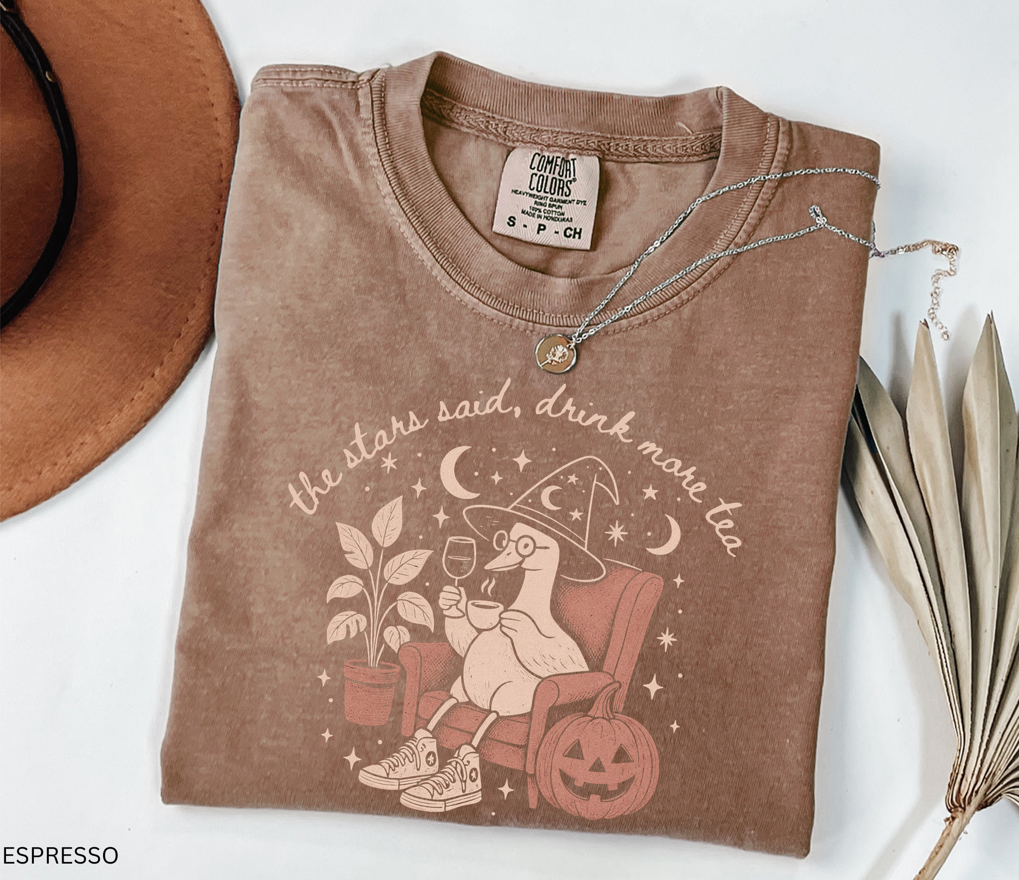 A folded espresso brown t-shirt with a cute cartoon duck wearing a witch hat and glasses, sitting in a pumpkin chair and holding a wine glass. The graphic includes the text 'the stars said, drink more tea' and is styled on a white background with a brown hat, a necklace, and dried palm leaves.