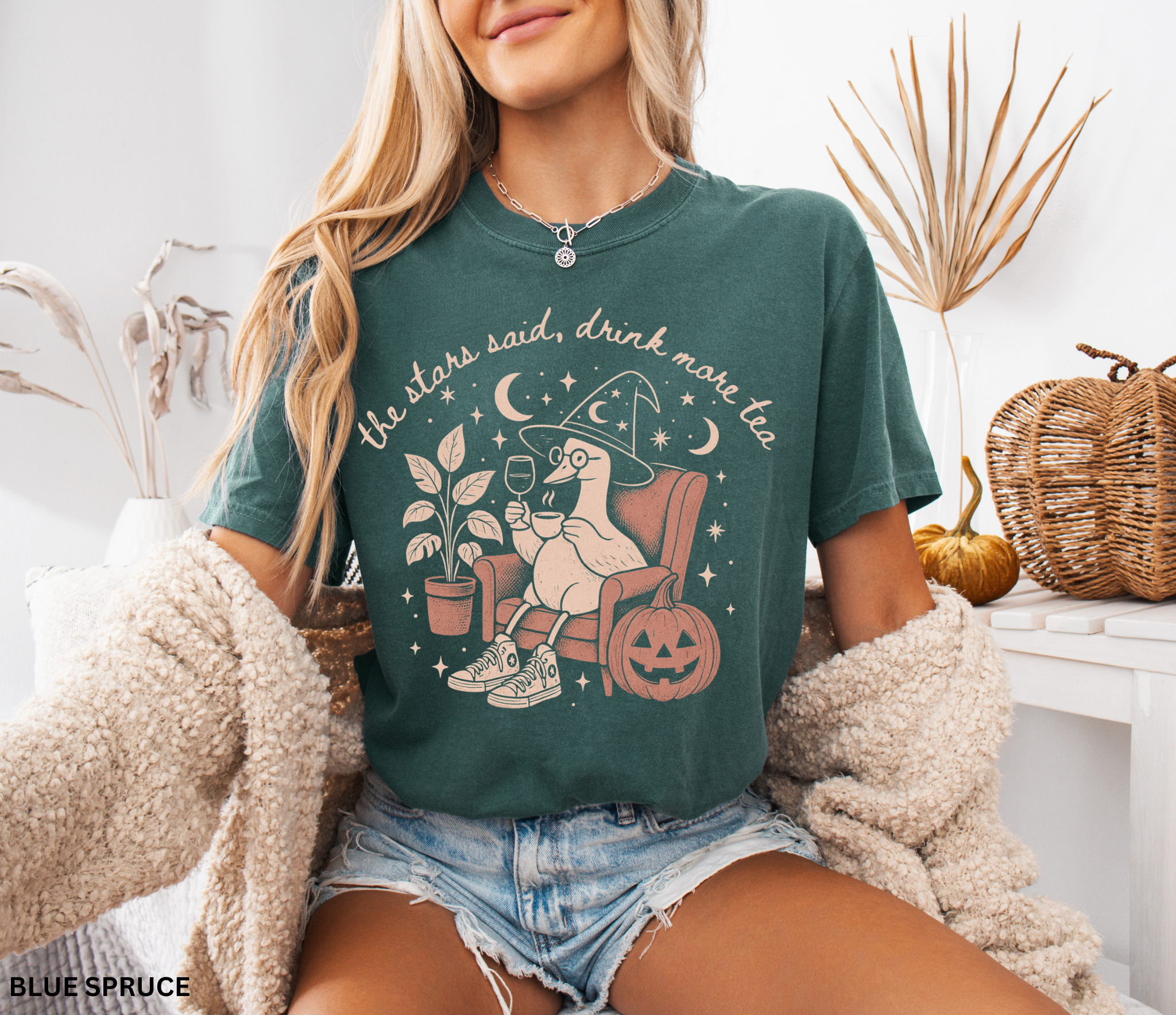 Comfort Colors graphic t-shirt featuring a cozy witch goose sitting in an armchair holding tea and wine beside a pumpkin with stars and moons and the quote "The Stars Said, Drink More Tea."