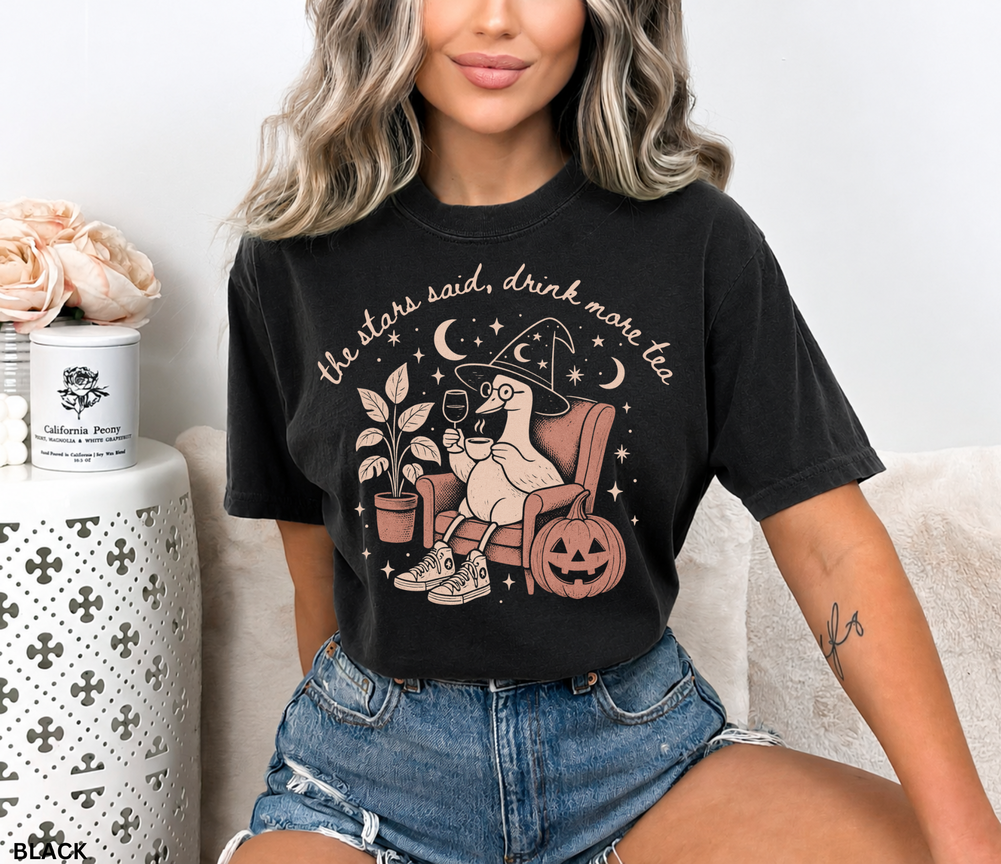 Comfort Colors graphic t-shirt featuring a cozy witch goose sitting in an armchair holding tea and wine beside a pumpkin with stars and moons and the quote "The Stars Said, Drink More Tea."