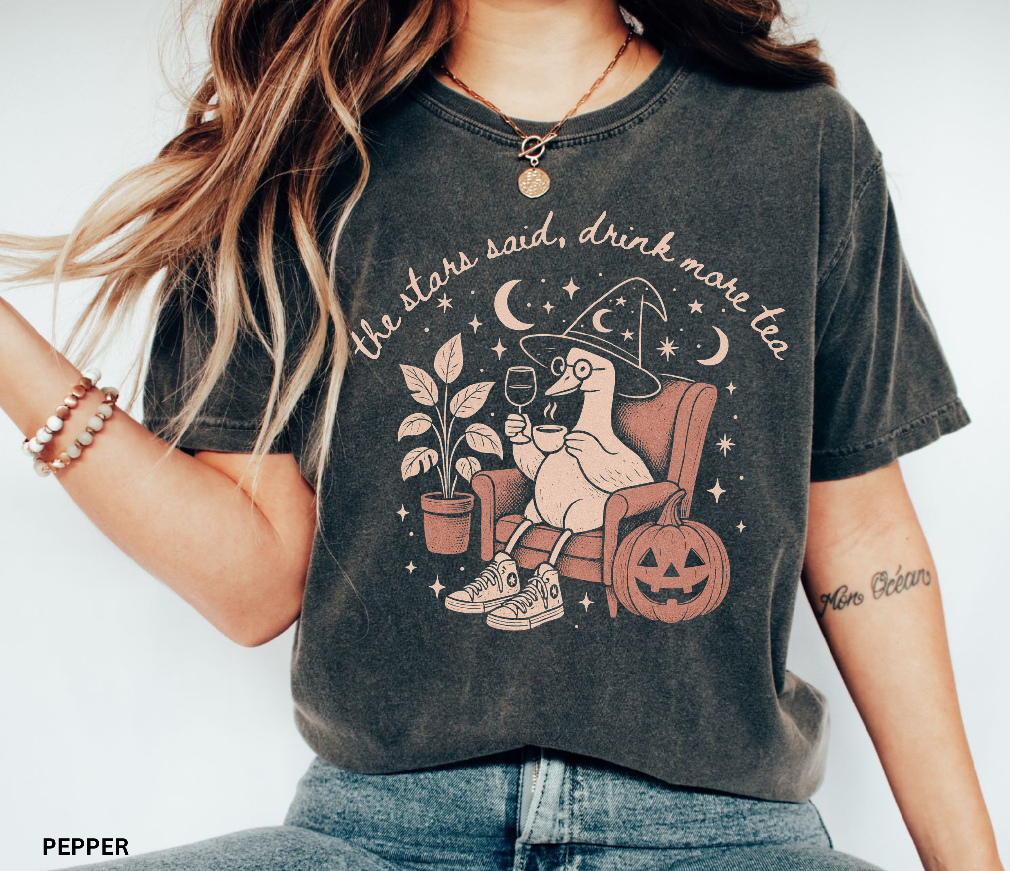 Comfort Colors graphic t-shirt featuring a cozy witch goose sitting in an armchair holding tea and wine beside a pumpkin with stars and moons and the quote "The Stars Said, Drink More Tea."