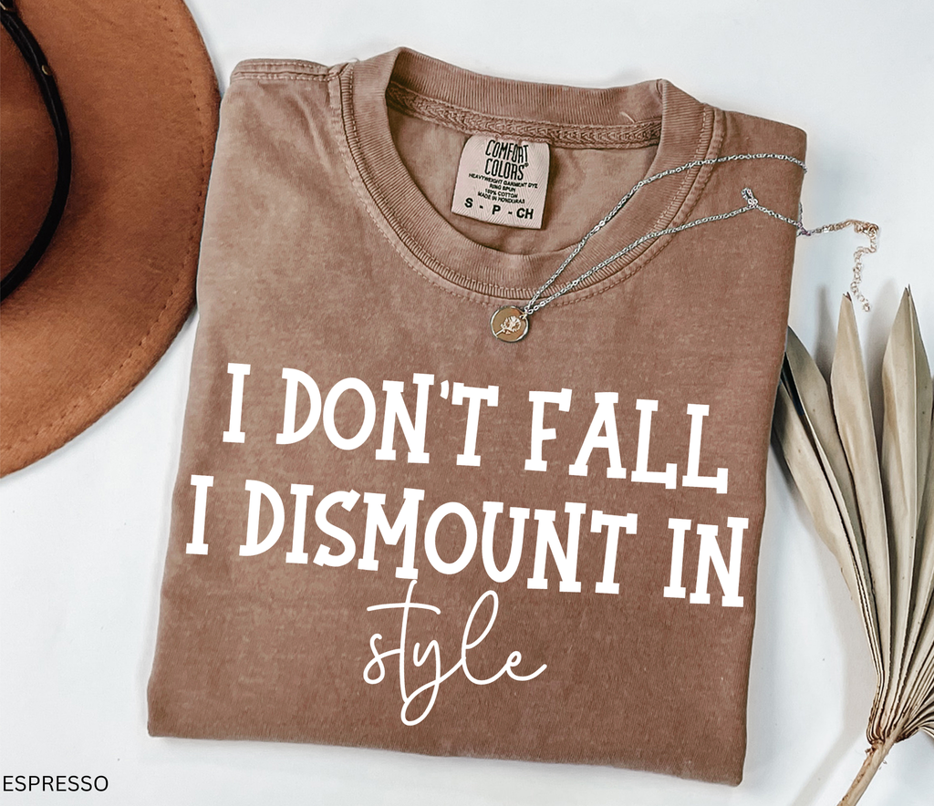 A folded espresso brown t-shirt with a white graphic print reading 'I DON'T FALL I DISMOUNT IN style' is styled in a flat-lay with a brown hat, a silver necklace, and dried palm leaves on a white background.
