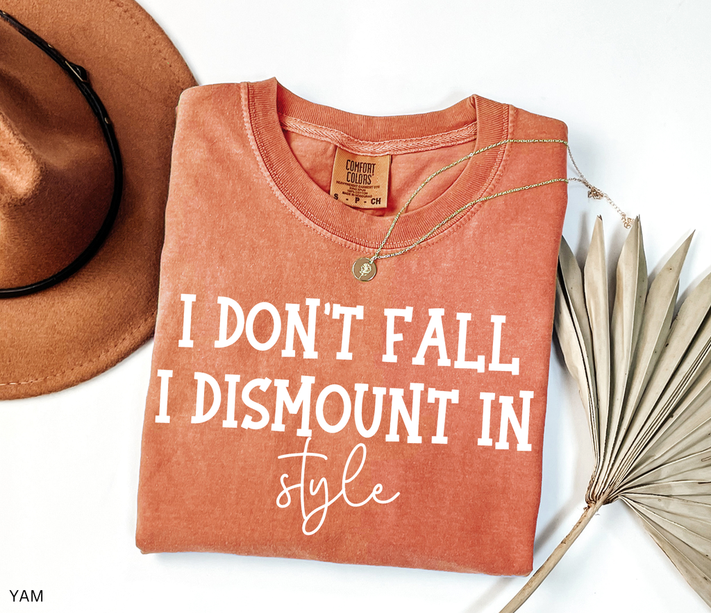 A folded terracotta-colored t-shirt with white text reading 'I DON'T FALL I DISMOUNT IN style' is styled in a flat lay with a brown hat, a gold necklace, and a dried palm leaf on a white background.