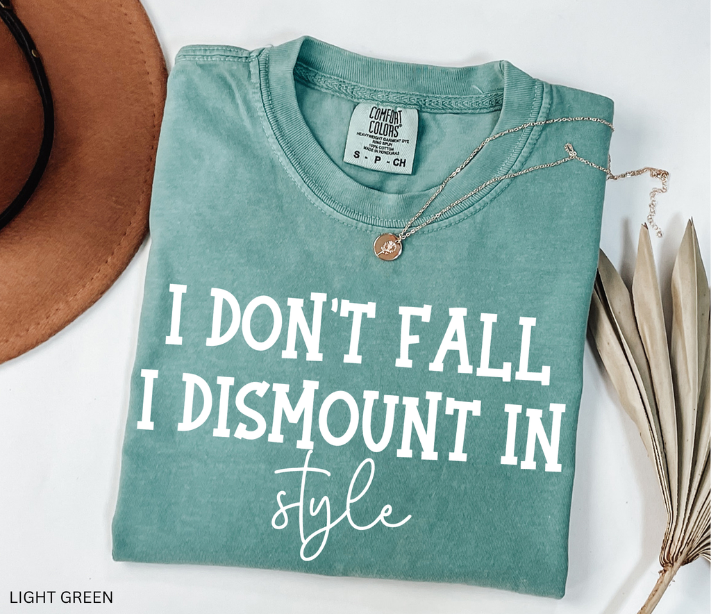 A folded light green graphic T-shirt with white text reading 'I DON'T FALL I DISMOUNT IN style' is styled on a white background with a tan hat edge, a necklace, and dried leaves. A small neck label reads Comfort Colors.