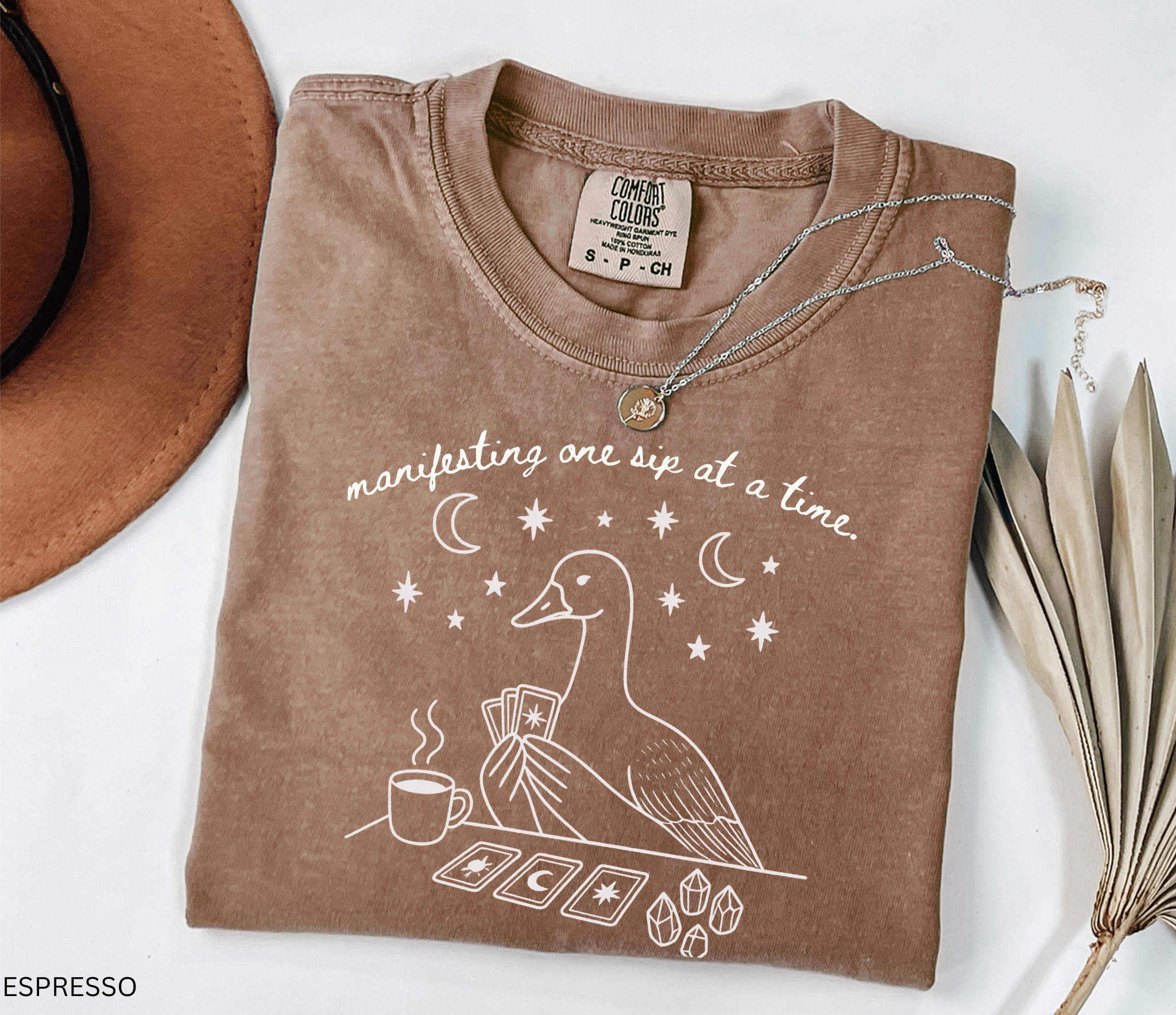 Comfort Colors t-shirt featuring a white tarot goose graphic with coffee, crystals, tarot cards, and the phrase "Manifesting One Sip at a Time."