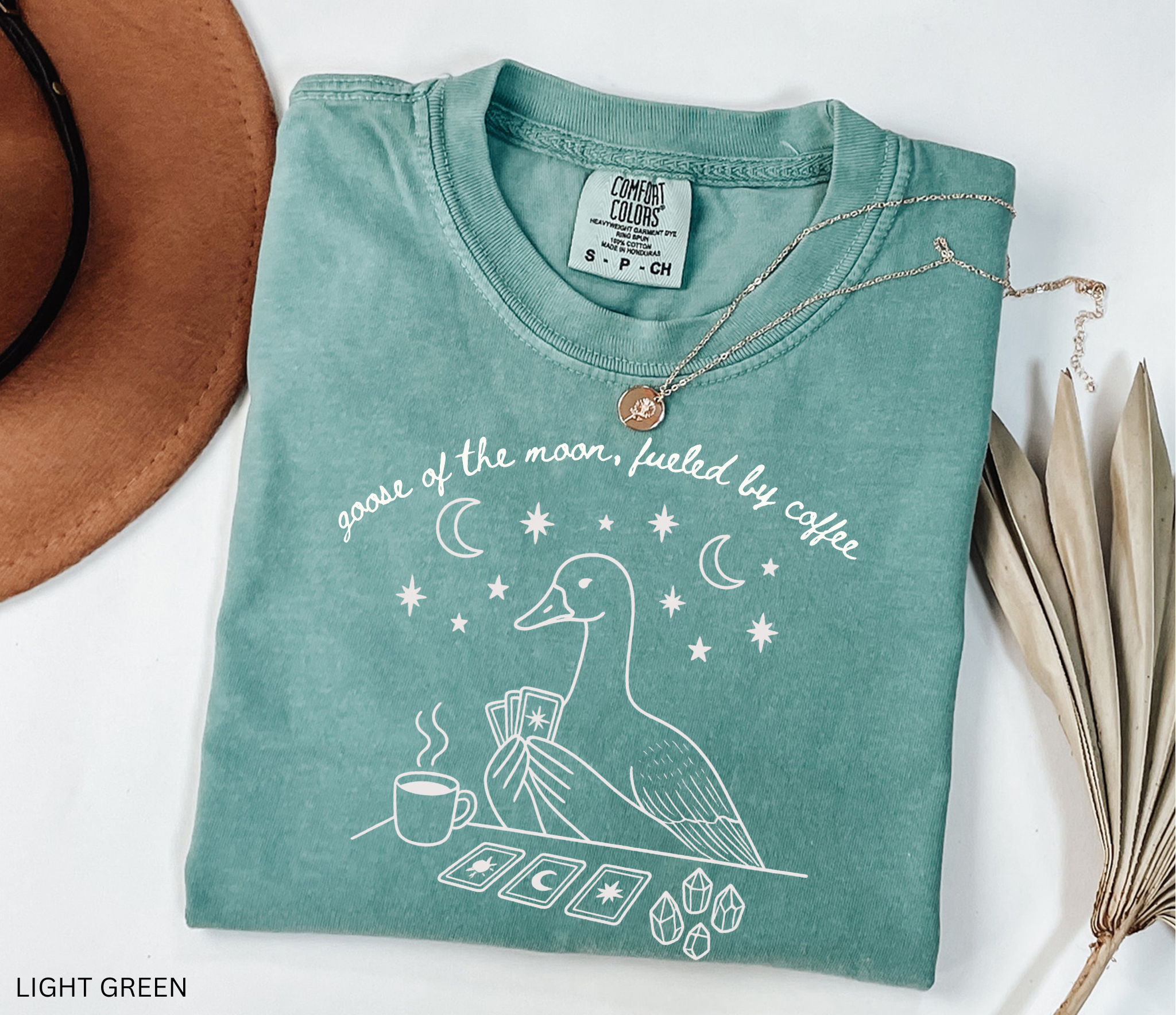 Comfort Colors t-shirt featuring a celestial goose with tarot cards, coffee cup, crystals, moons and stars and the phrase "Goose of the Moon, Fueled by Coffee."