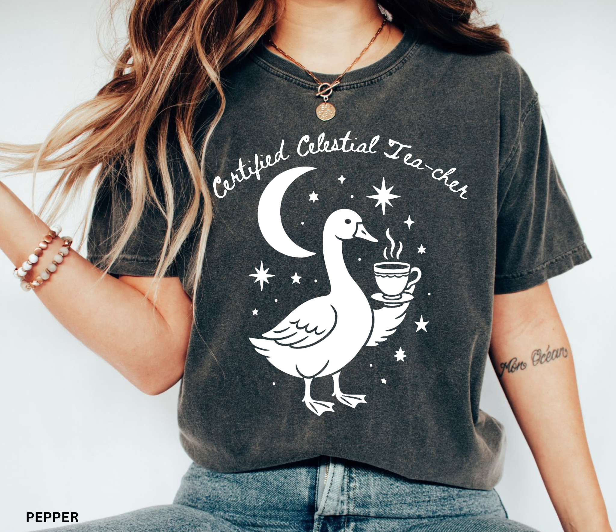 A woman wearing a dark charcoal gray washed graphic t-shirt with a white print of a goose, crescent moon, stars, and the text 'Certified Celestial Tea-cher'.