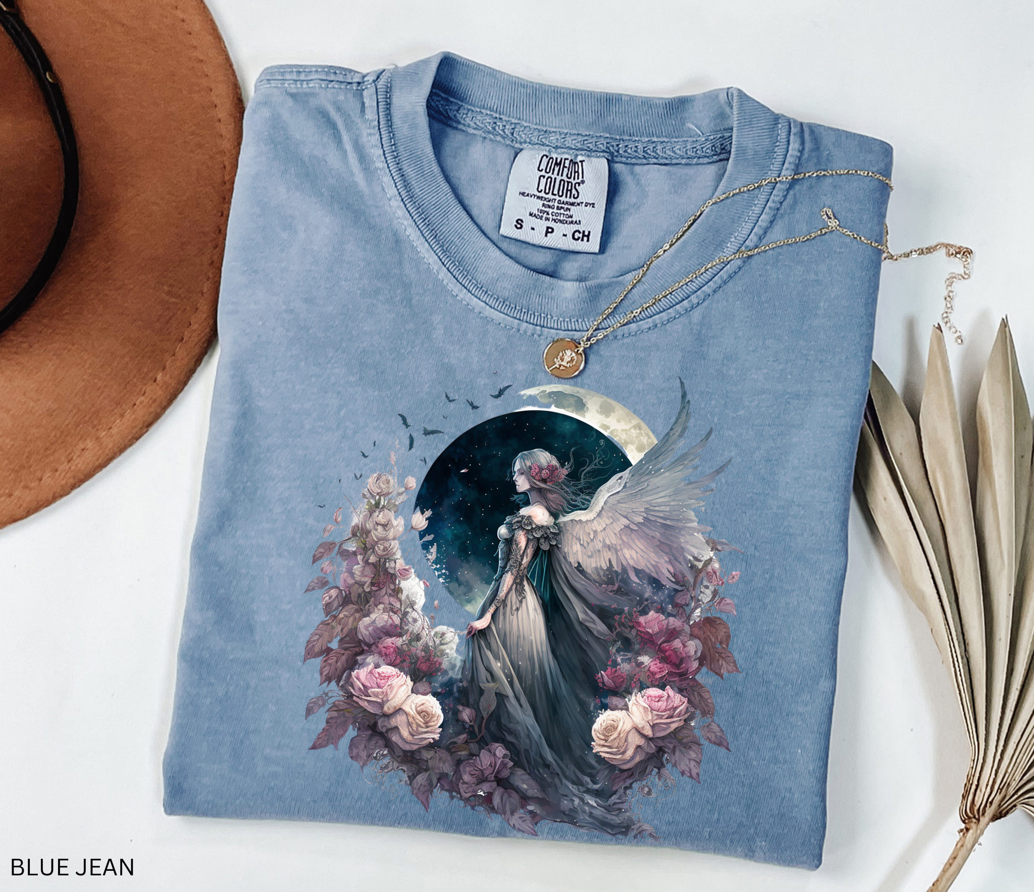 A folded light blue denim T-shirt with a detailed fantasy graphic of an angel surrounded by pink and purple flowers under a crescent moon. The shirt is styled in a flat-lay with a gold necklace, a brown hat, and dried palm leaves on a white background.
