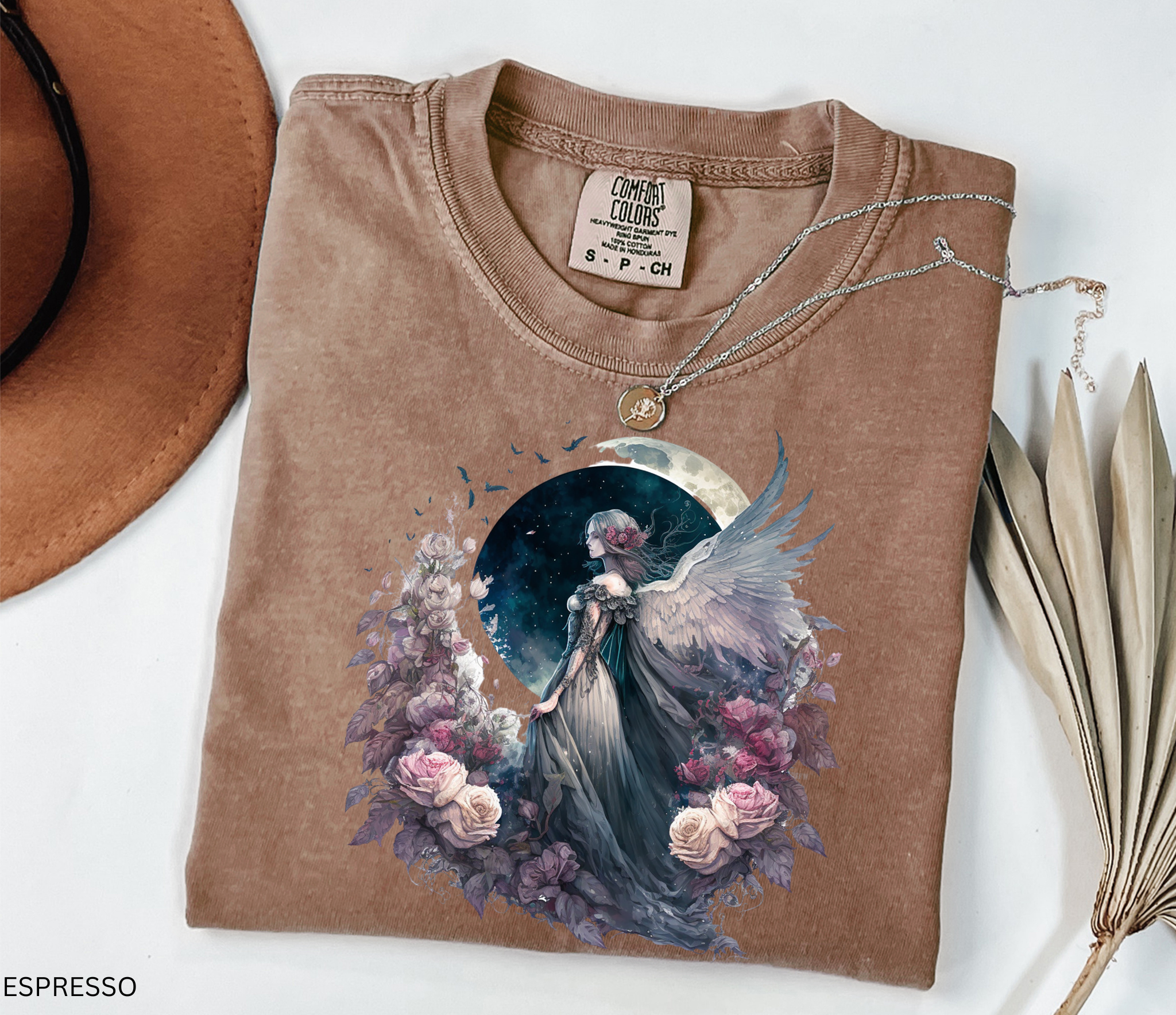 A folded espresso brown t-shirt with a detailed graphic of an angel in a blue dress surrounded by pink and purple flowers under a crescent moon, styled with a necklace, a hat, and dried leaves on a white background.