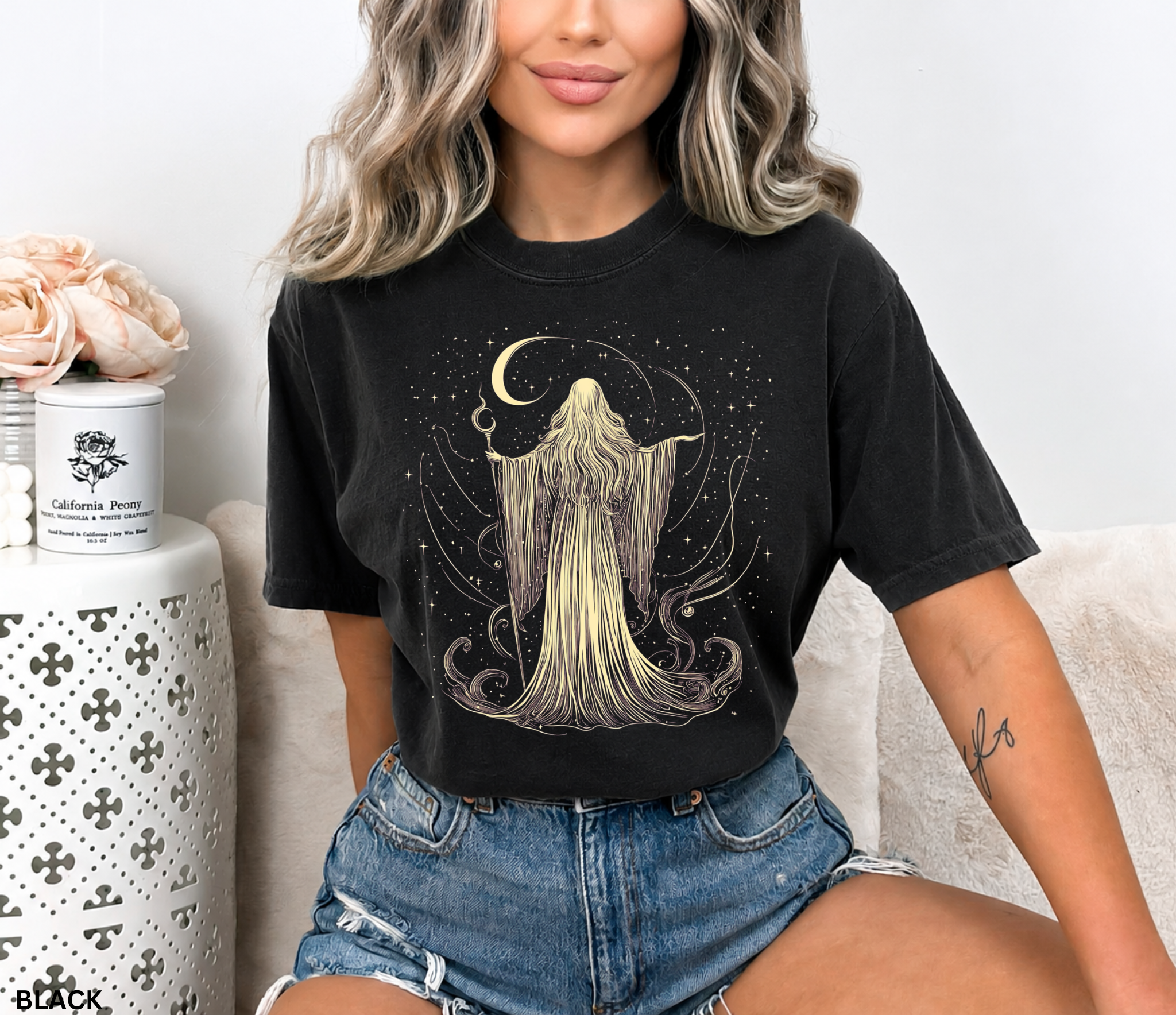 A woman wearing a black graphic t-shirt with a cream-colored illustration of a hooded figure under a crescent moon, paired with denim shorts, sitting on a couch.