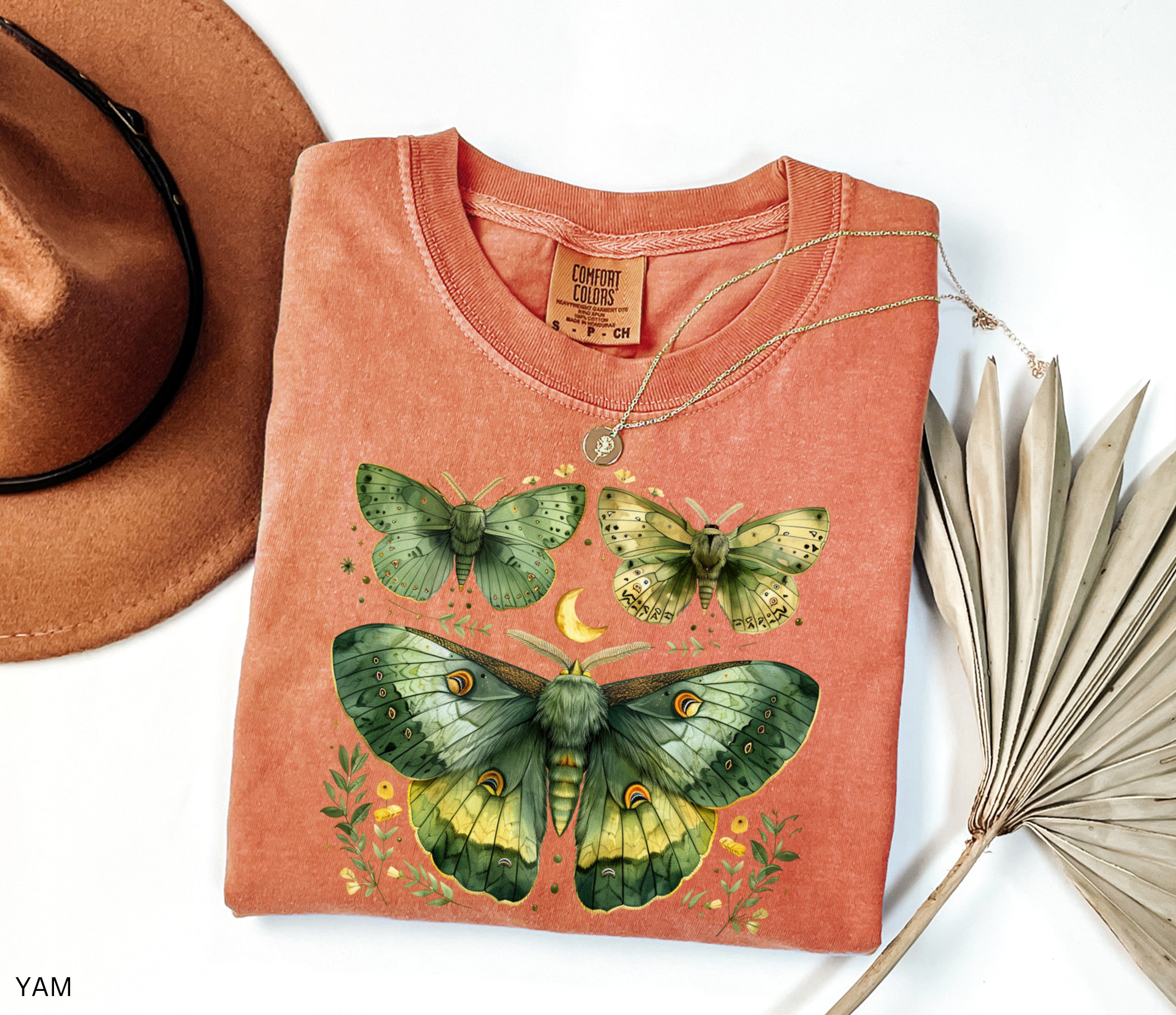 A folded terracotta-colored t-shirt with a detailed green and yellow moth and butterfly graphic, styled in a flat lay with a brown hat, a gold necklace, and dried palm leaves on a white background.