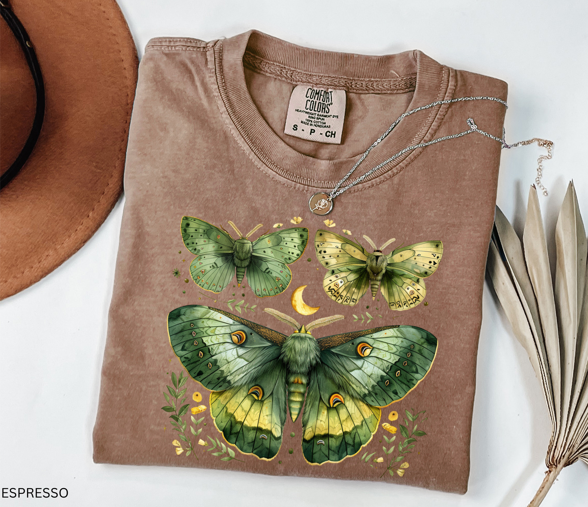A folded espresso brown t-shirt with a detailed graphic of three green and yellow moths and a crescent moon, styled on a white background with a brown hat, a silver necklace, and dried leaves.