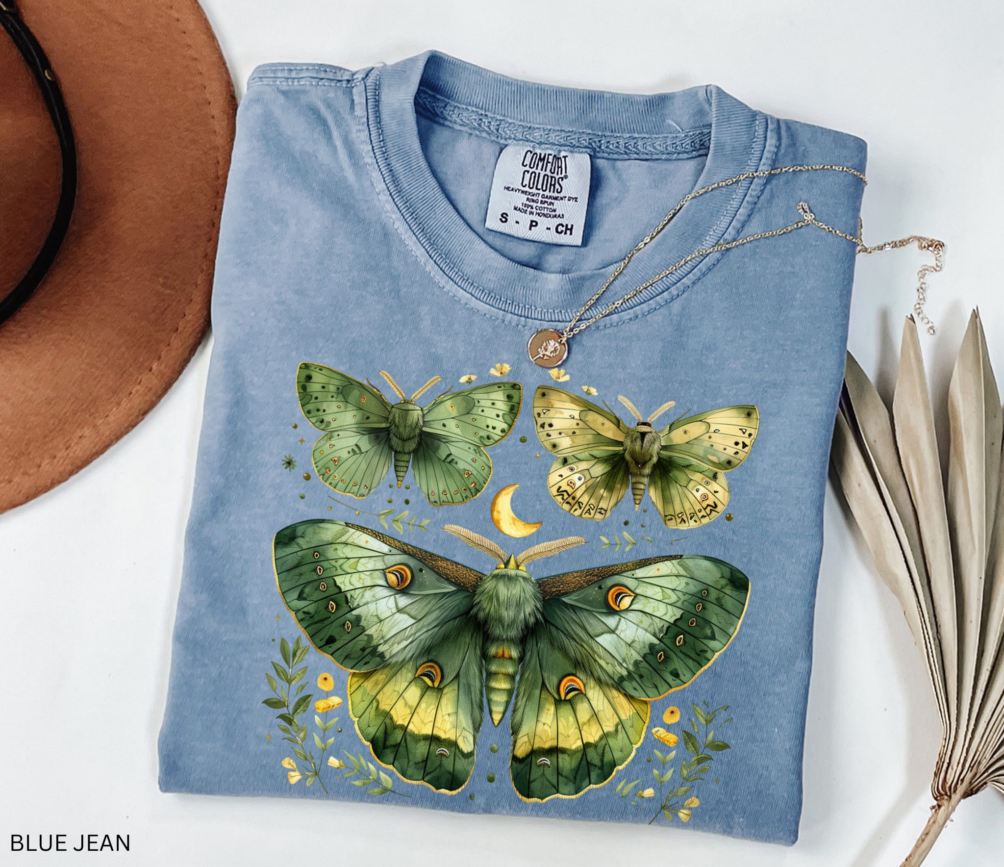 A folded light blue t-shirt with a detailed graphic of green and yellow moths and butterflies, styled with a gold necklace on a white background with a brown hat and dried palm leaves.