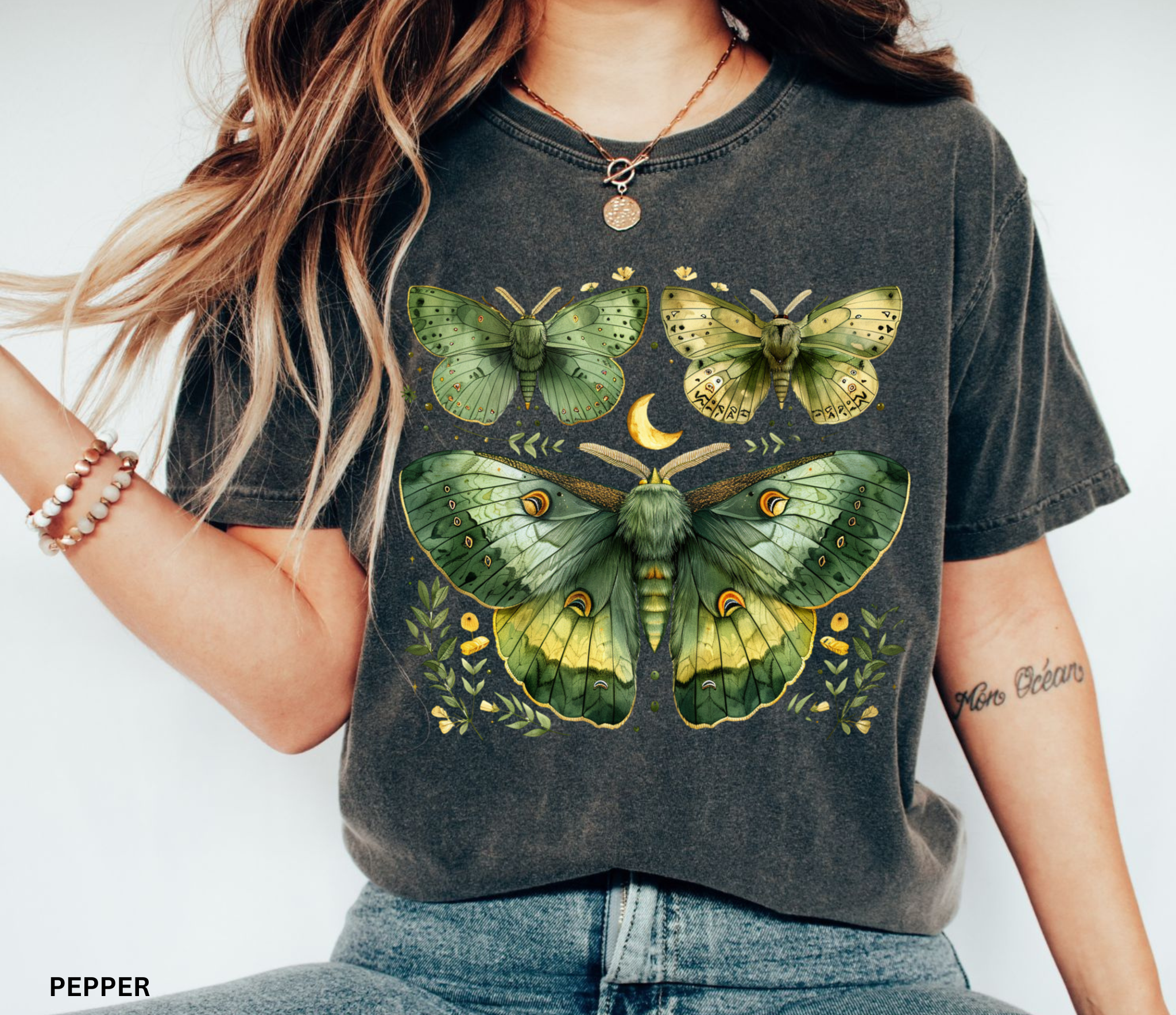 A woman wearing a dark charcoal gray graphic t-shirt with a large green and yellow moth and butterfly print on the front, paired with blue jeans. The text 'PEPPER' is visible in the bottom-left corner.