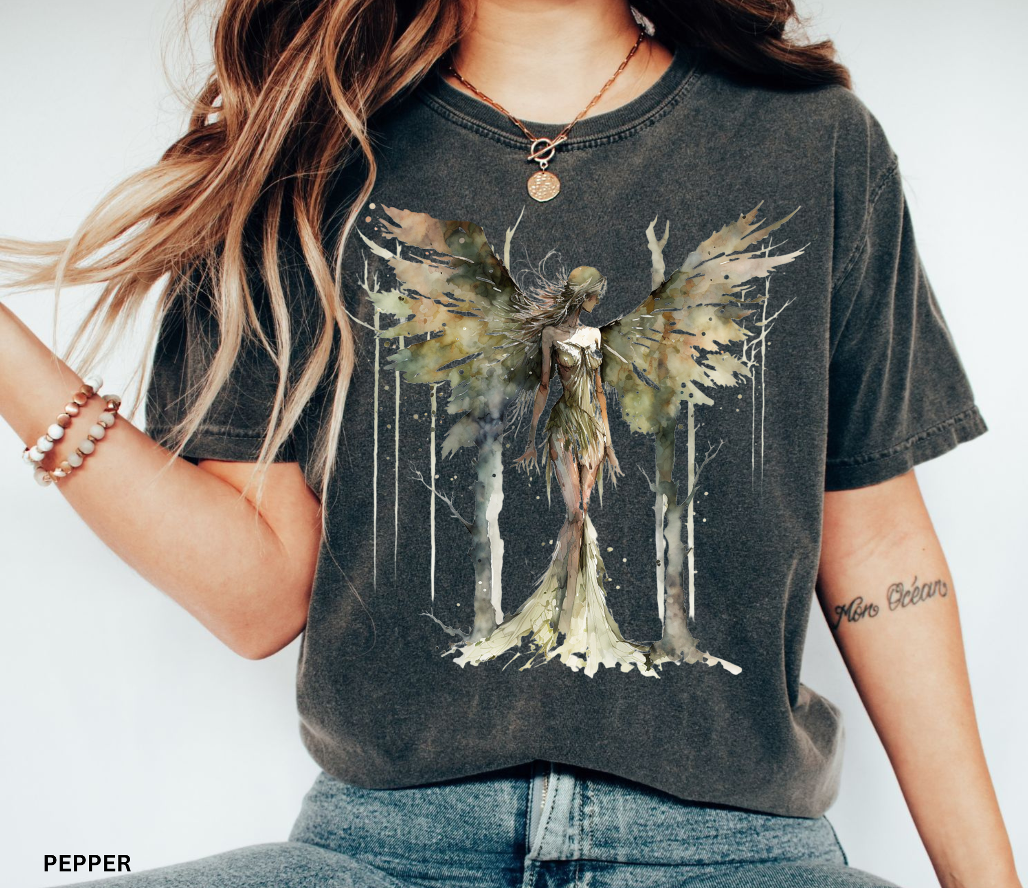 A woman wearing a dark charcoal gray graphic t-shirt with a watercolor-style winged fairy design on the front, paired with blue jeans. The text 'PEPPER' is visible in the bottom-left corner.
