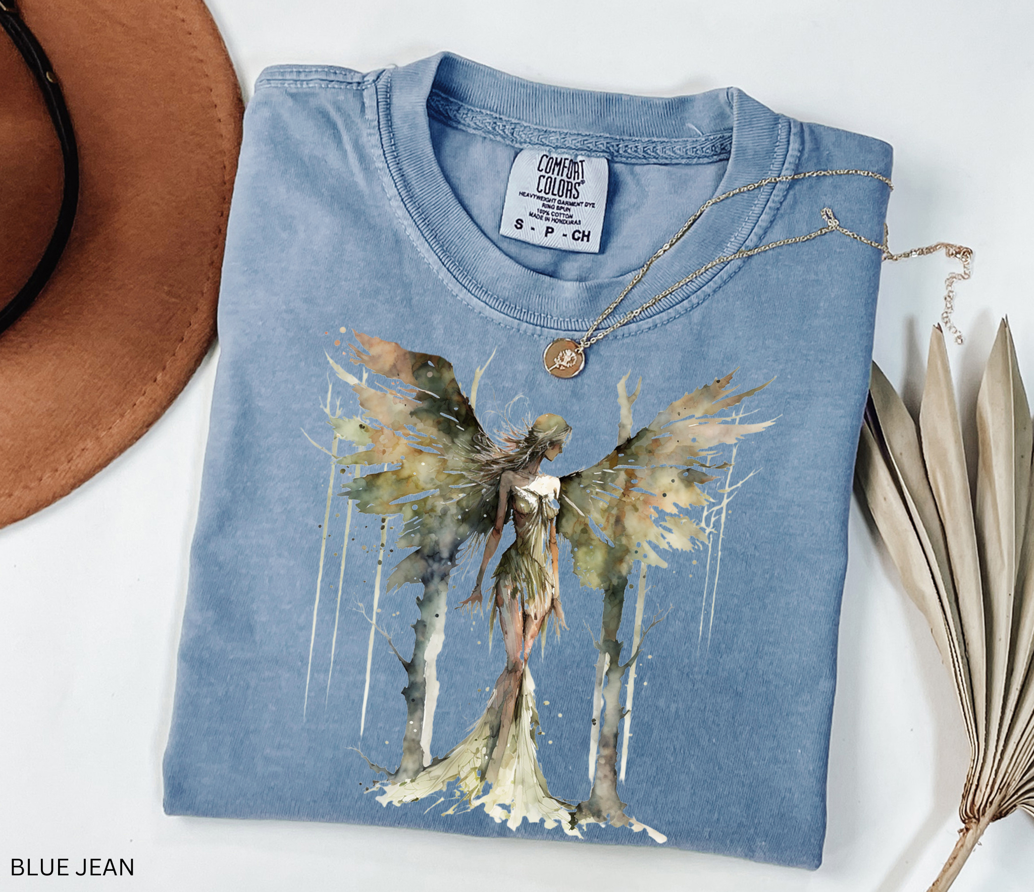 A folded light blue t-shirt with a watercolor-style angel graphic on the front, styled in a flat lay with a brown hat, a gold necklace, and dried palm leaves on a white background.