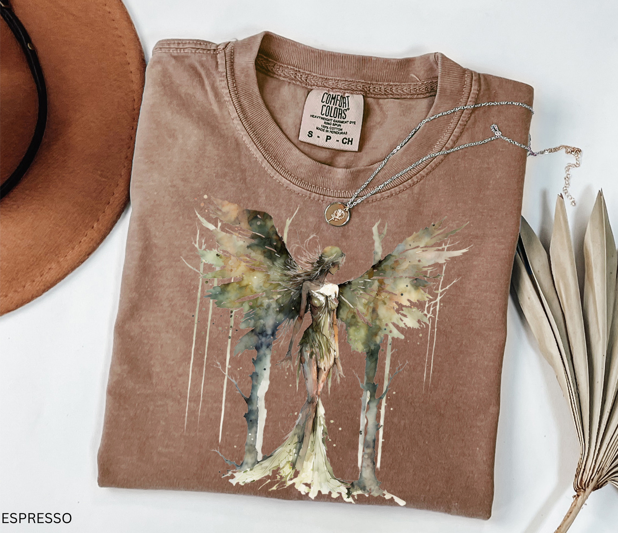 A folded espresso brown t-shirt with a watercolor-style winged fairy graphic on the front, styled flat-lay with a brown hat, dried palm leaves, and a silver necklace on a white background.