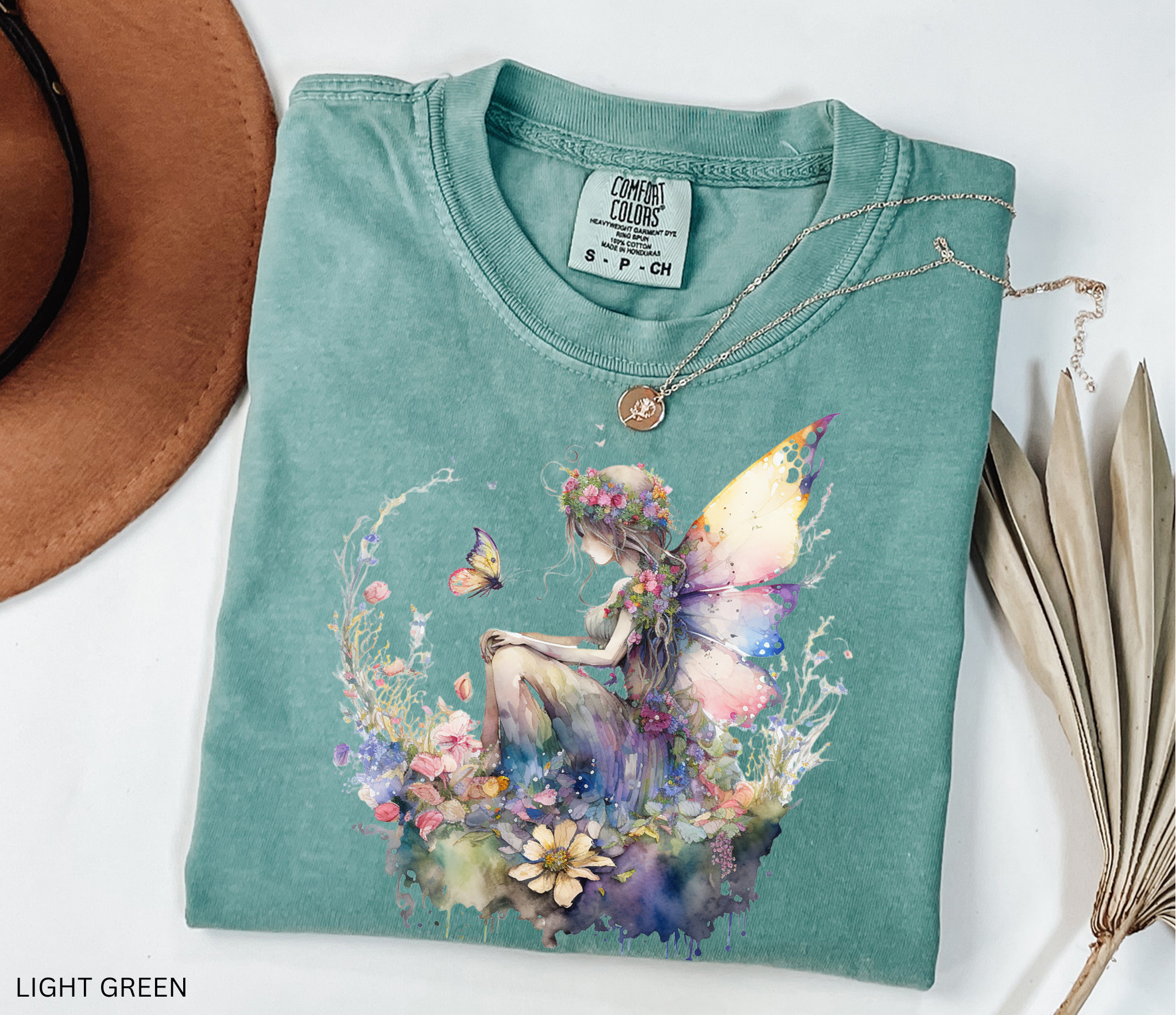 A folded light green t-shirt with a colorful fairy and floral graphic print, styled with a gold necklace, a brown hat, and dried leaves on a white background.