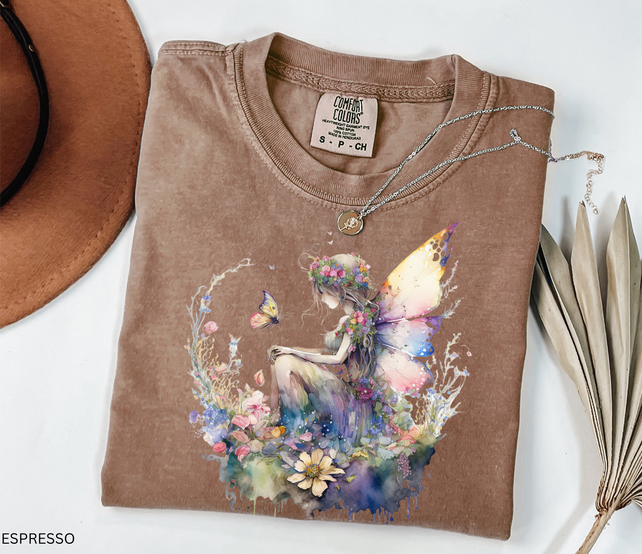 An espresso brown folded t-shirt with a colorful watercolor fairy graphic, styled with a necklace, a brown hat, and dried leaves on a white background.
