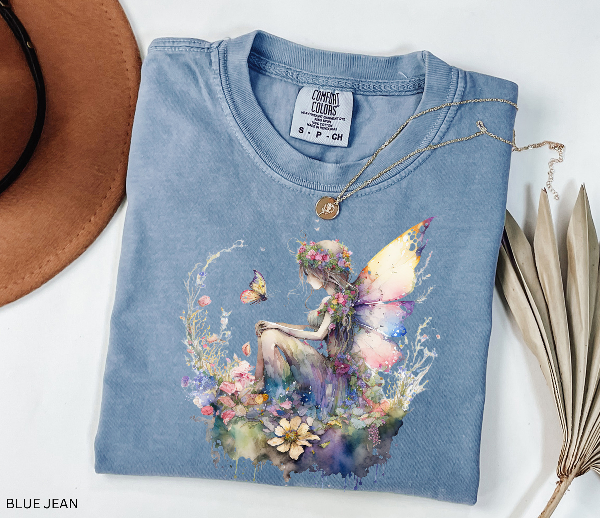 A folded light blue t-shirt with a colorful watercolor fairy graphic on the front, styled on a white background with a brown hat, a gold necklace, and dried leaves. The text 'BLUE JEAN' is visible in the bottom-left corner.