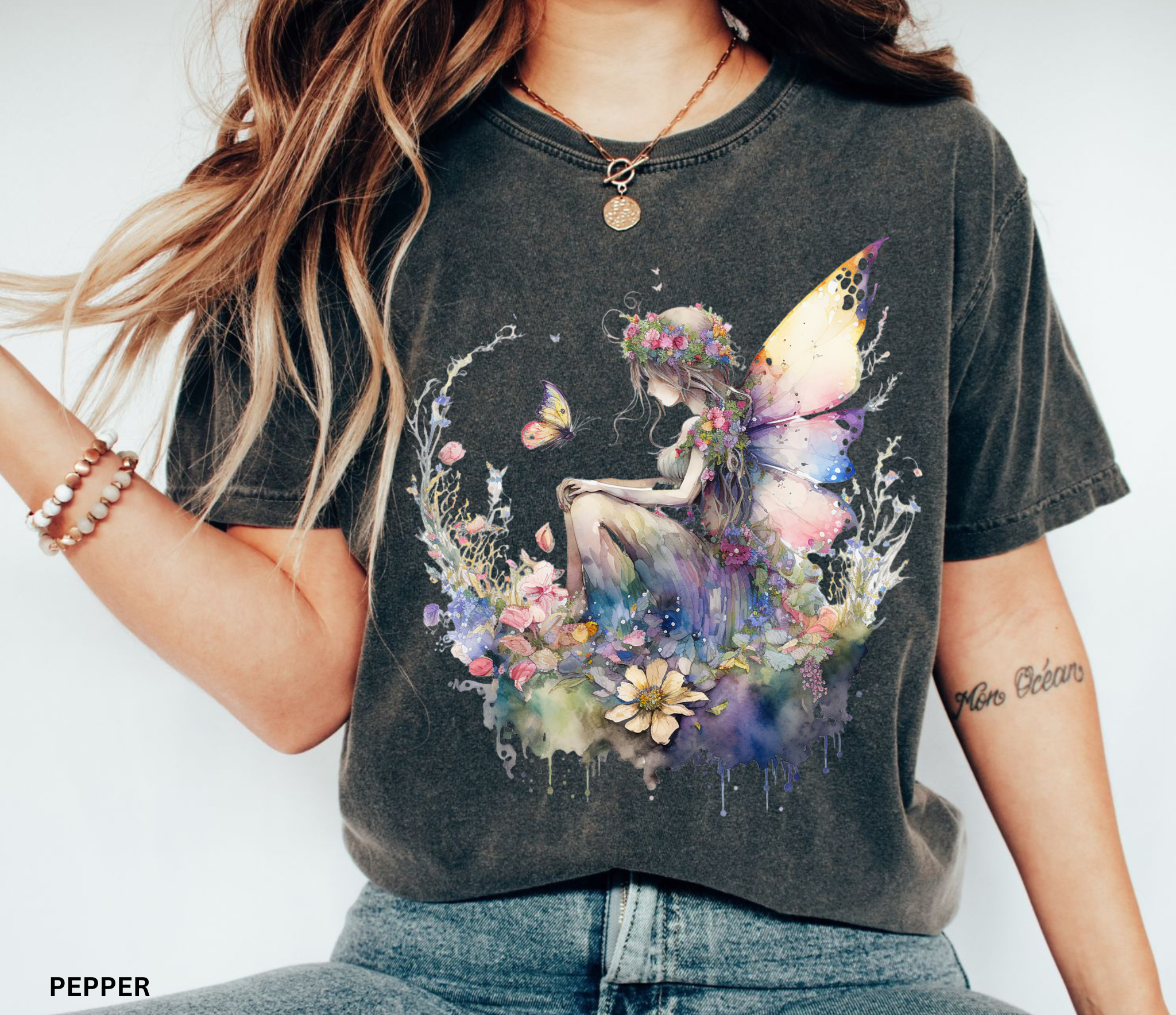 A woman wearing a dark charcoal gray graphic t-shirt with a colorful watercolor design of a fairy surrounded by flowers. The text 'PEPPER' is visible in the bottom-left corner, indicating the shirt's colorway.