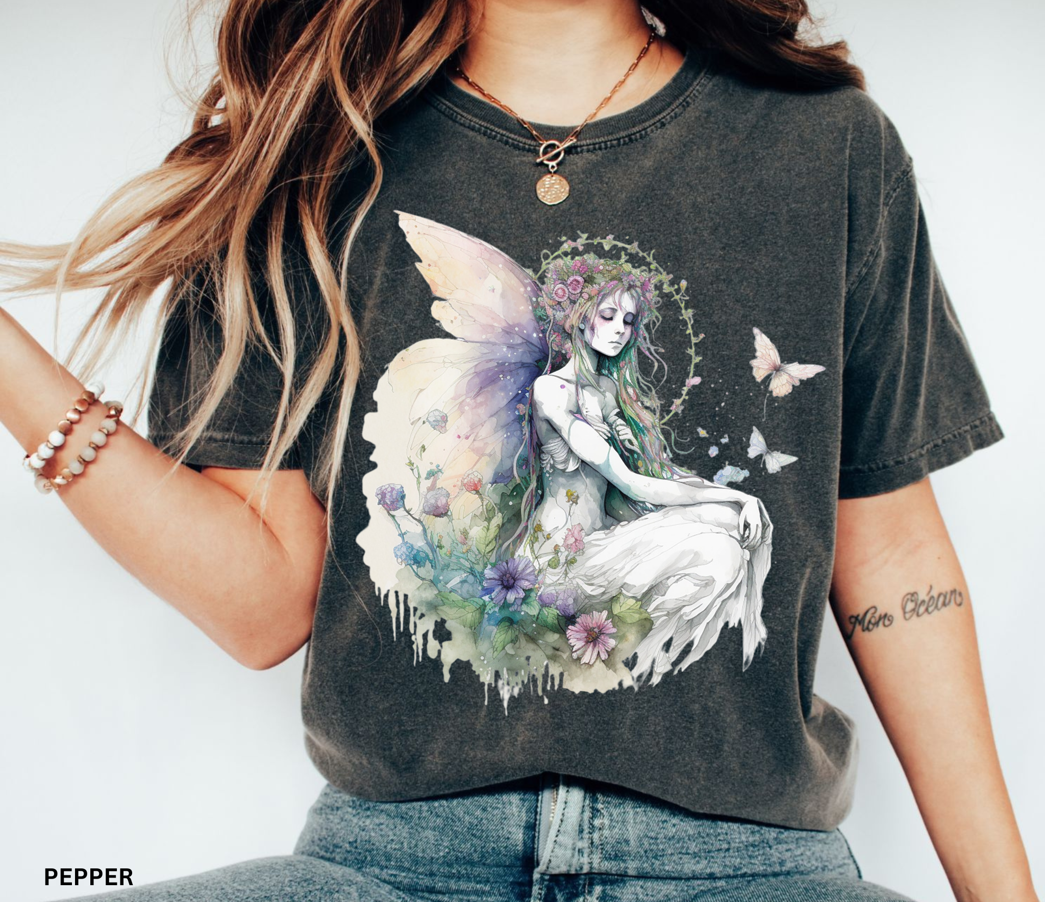 A woman wearing a dark charcoal gray graphic t-shirt with a detailed illustration of a fairy in a white dress surrounded by flowers and butterflies. The text 'PEPPER' is visible in the bottom-left corner, indicating the colorway.