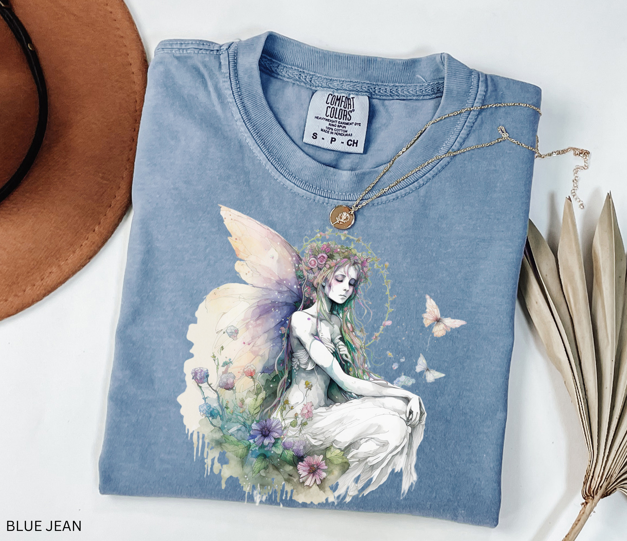 A folded light blue t-shirt with a detailed fantasy graphic of a winged fairy surrounded by flowers and butterflies. The shirt is styled on a white background with a brown hat corner, a gold necklace, and dried leaves, with 'BLUE JEAN' text in the bottom-left corner.
