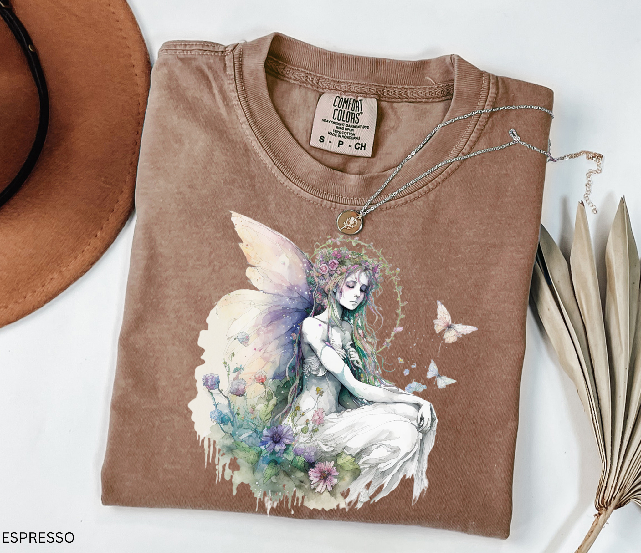 A folded espresso brown t-shirt with a detailed fantasy graphic of a winged fairy surrounded by flowers and butterflies, styled with a necklace, a brown hat, and dried leaves on a white background.